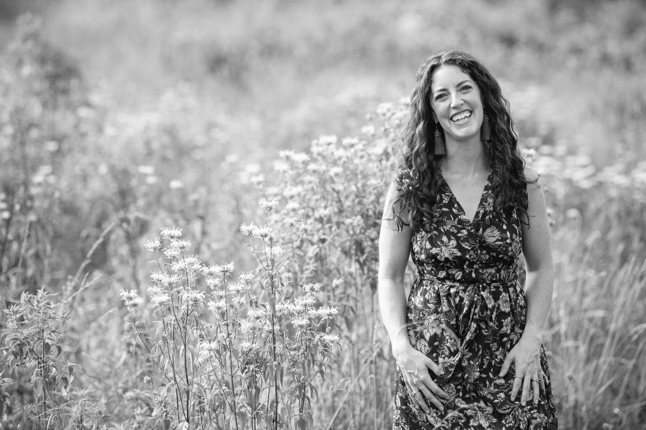 A smiling woman in a floral dress standing in a field of wildflowers, captured in black and white.