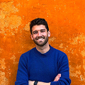 A smiling man with a beard and short hair, wearing a blue shirt, standing against an orange background.
