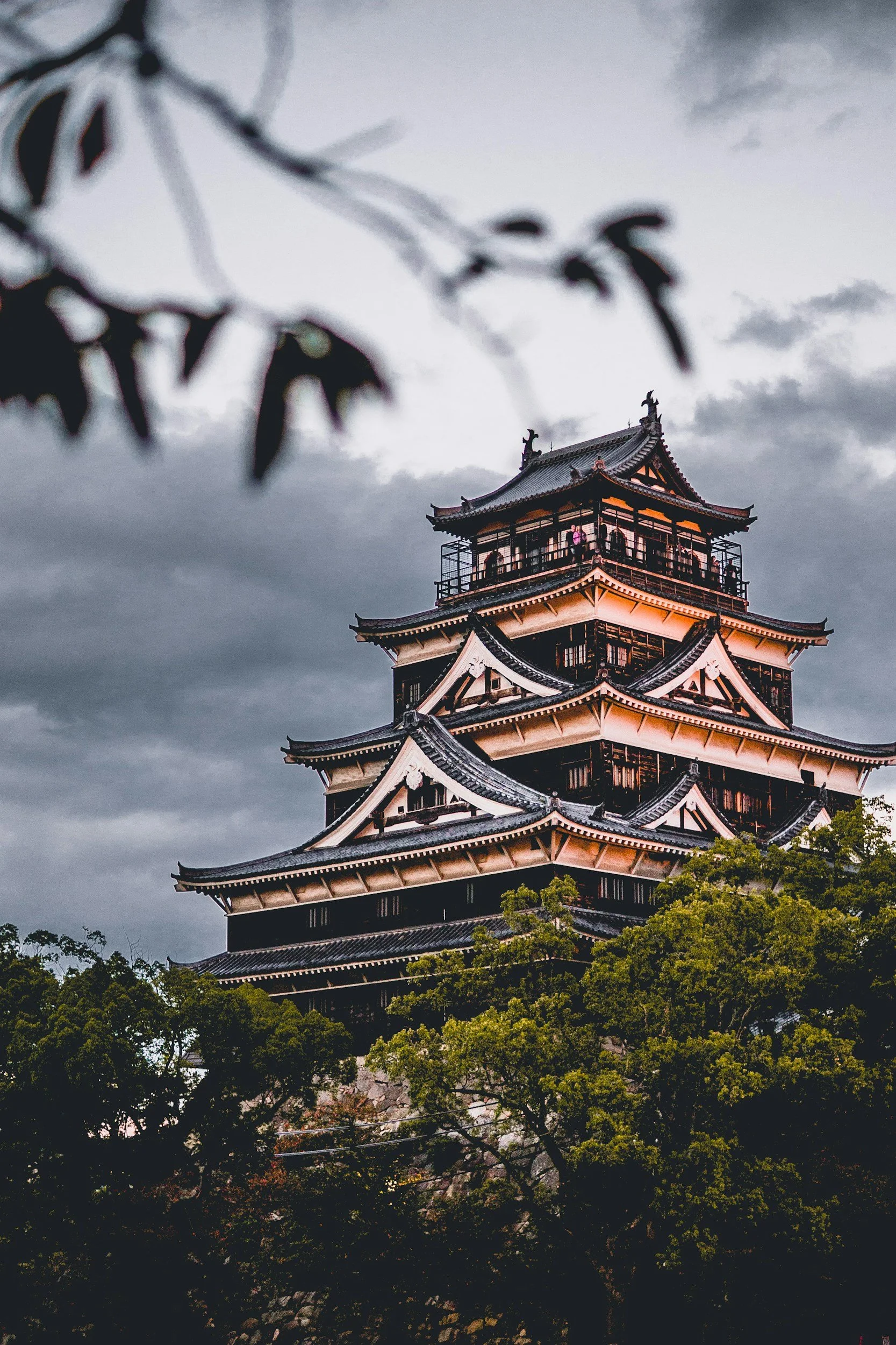 A traditional Japanese castle with multiple rooftops, framed by tree branches in the foreground, under a cloudy sky at dusk.