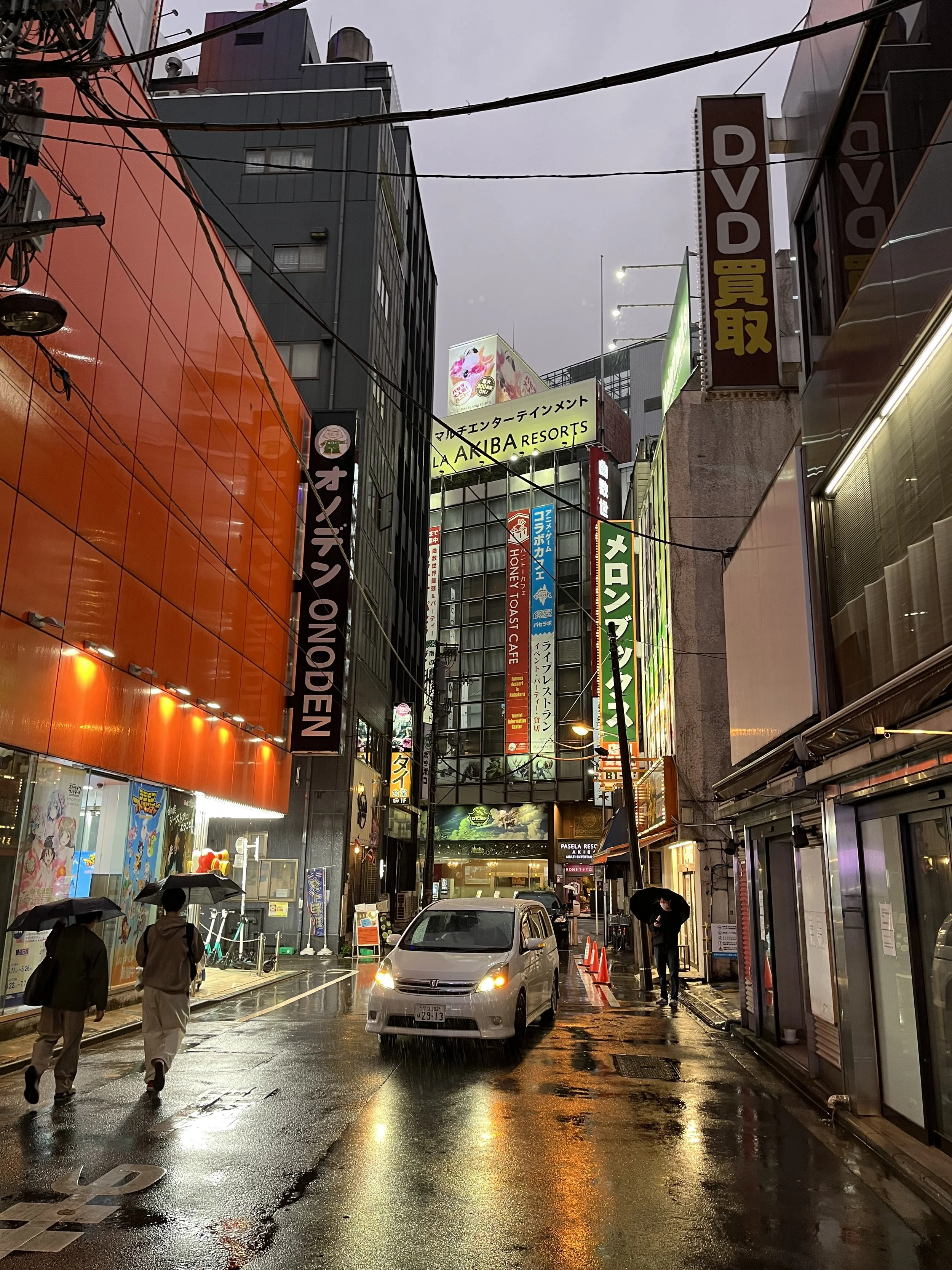 A rainy city street scene at dusk with illuminated signs in Japanese and English, two people walking with umbrellas, and a car on a wet road.