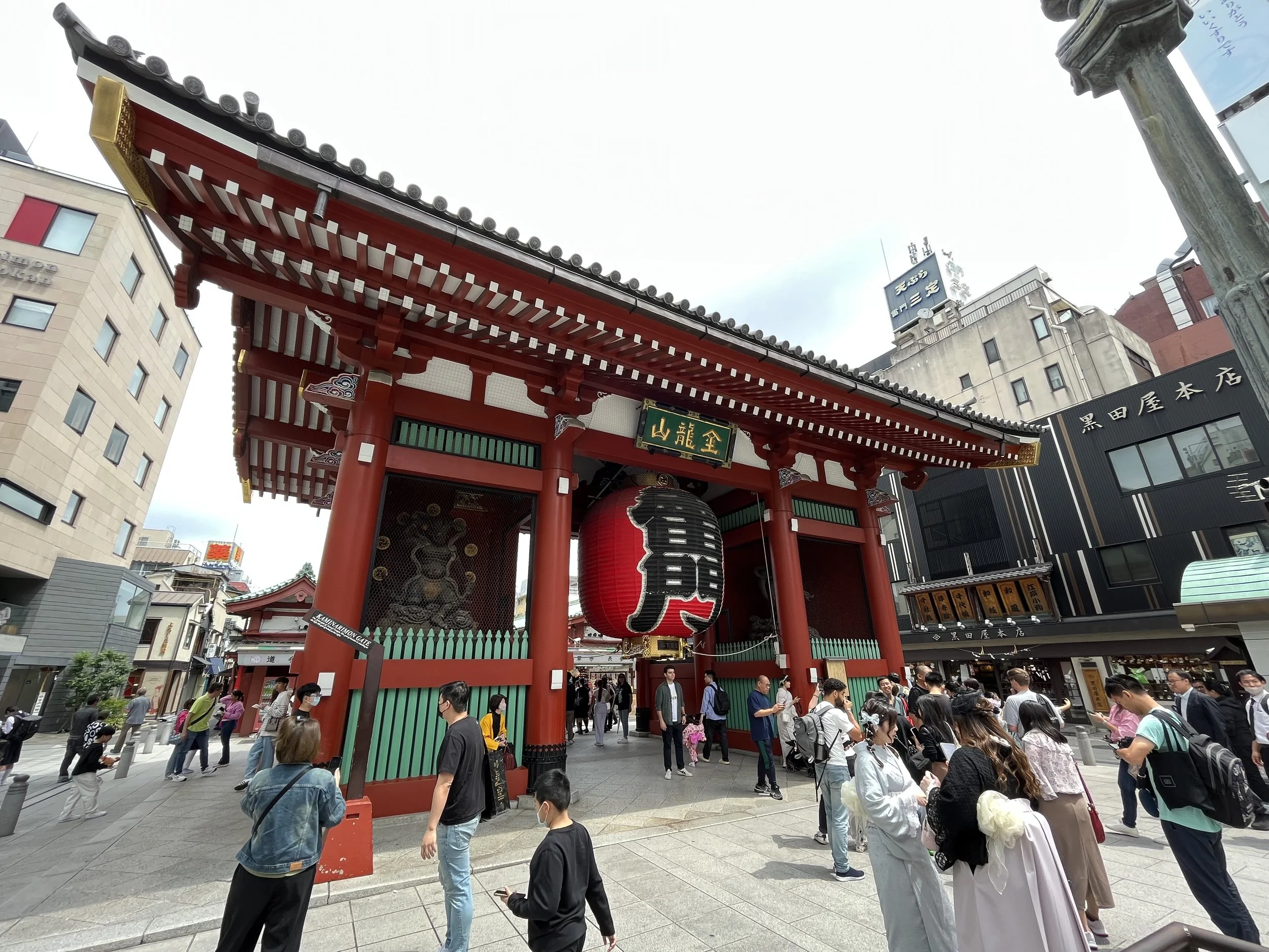 Entrance gate to a Japanese temple with a large red paper lantern, red and white wooden structure, and people walking and taking photos in front.