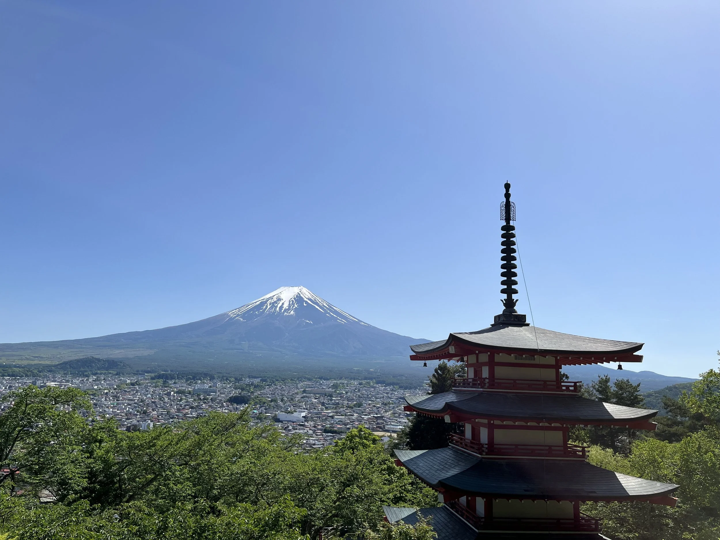 A view of Mount Fuji with snow on top, a traditional Japanese pagoda in the foreground, and a city below. Clear blue sky.