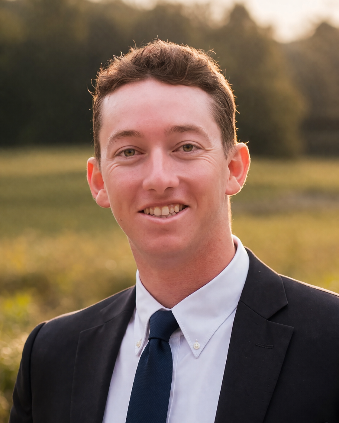 A young man in a suit smiling outdoors during golden hour with a blurred natural background of trees and greenery.