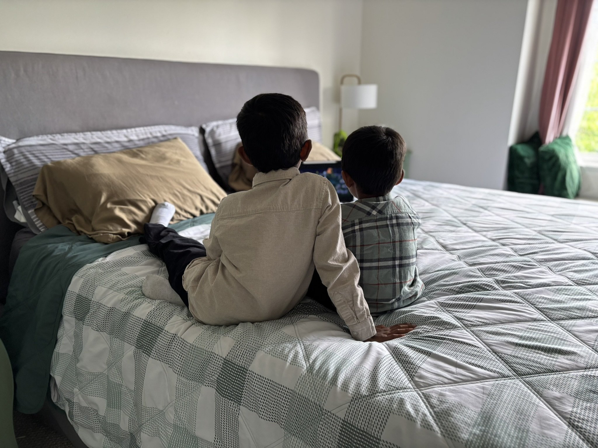 Two boys sitting on a bed, watching a tablet or phone in a bright bedroom. The bed has a gray headboard, beige and striped pillows, and a patterned quilt. There is a white nightstand and lamp beside the bed, with a window letting in natural light.