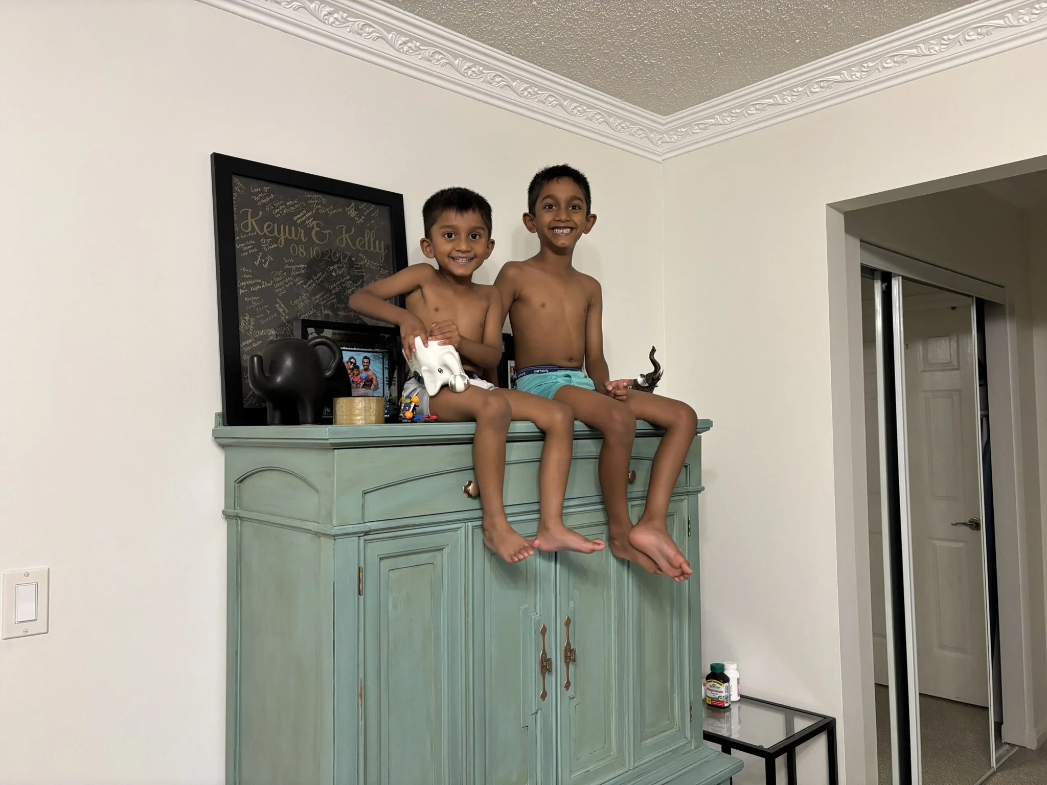 Two young boys sitting on top of a light green cabinet in a room, smiling at the camera. The boy on the left holds a white toy and the boy on the right holds a silver toy. Behind them is a black frame with a mirror and signatures, a photo, and a black figurine. The room has beige walls, a decorative ceiling, and a small black table with some bottles on it.