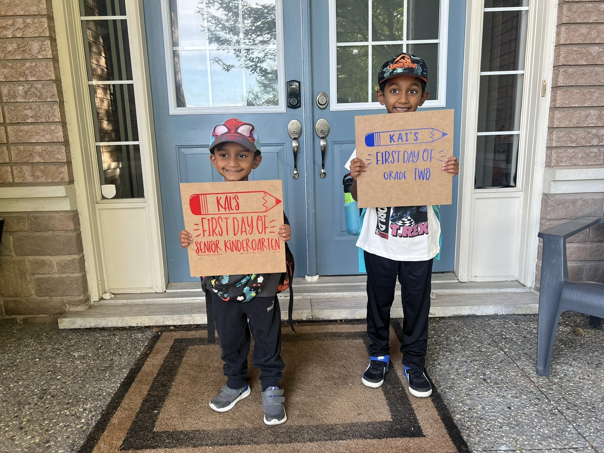 Two young boys standing on a doormat in front of a blue door, holding signs that read "Kai's First Day of Senior Kindergarten" and "Kai's First Day of Grade Two," both smiling.