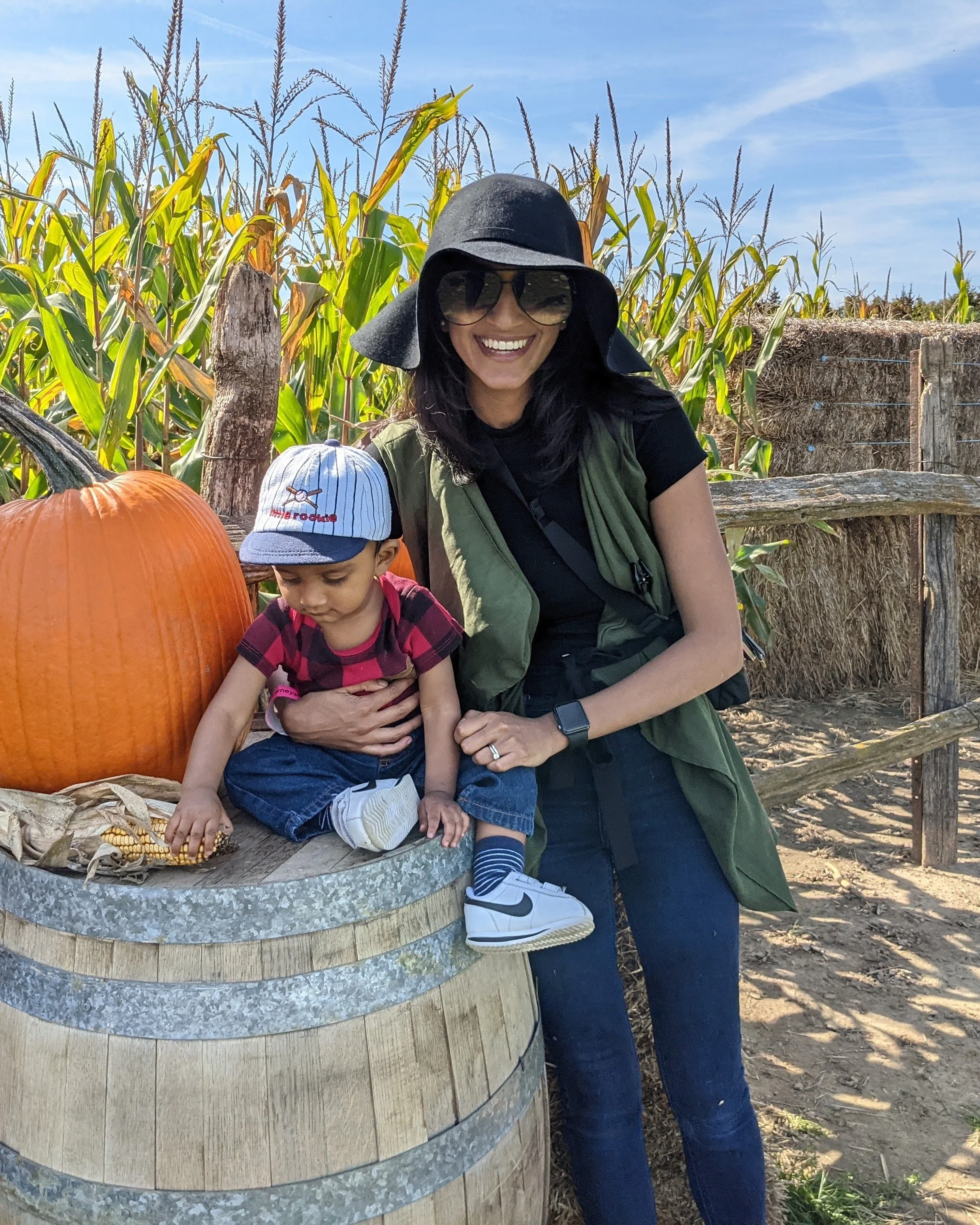 A woman and a young boy at a pumpkin patch, with tall corn stalks in the background. The woman is smiling, wearing sunglasses, a wide-brimmed hat, and a green vest, while the boy is focused on an ear of corn on a barrel, wearing a striped hat and a red plaid shirt.