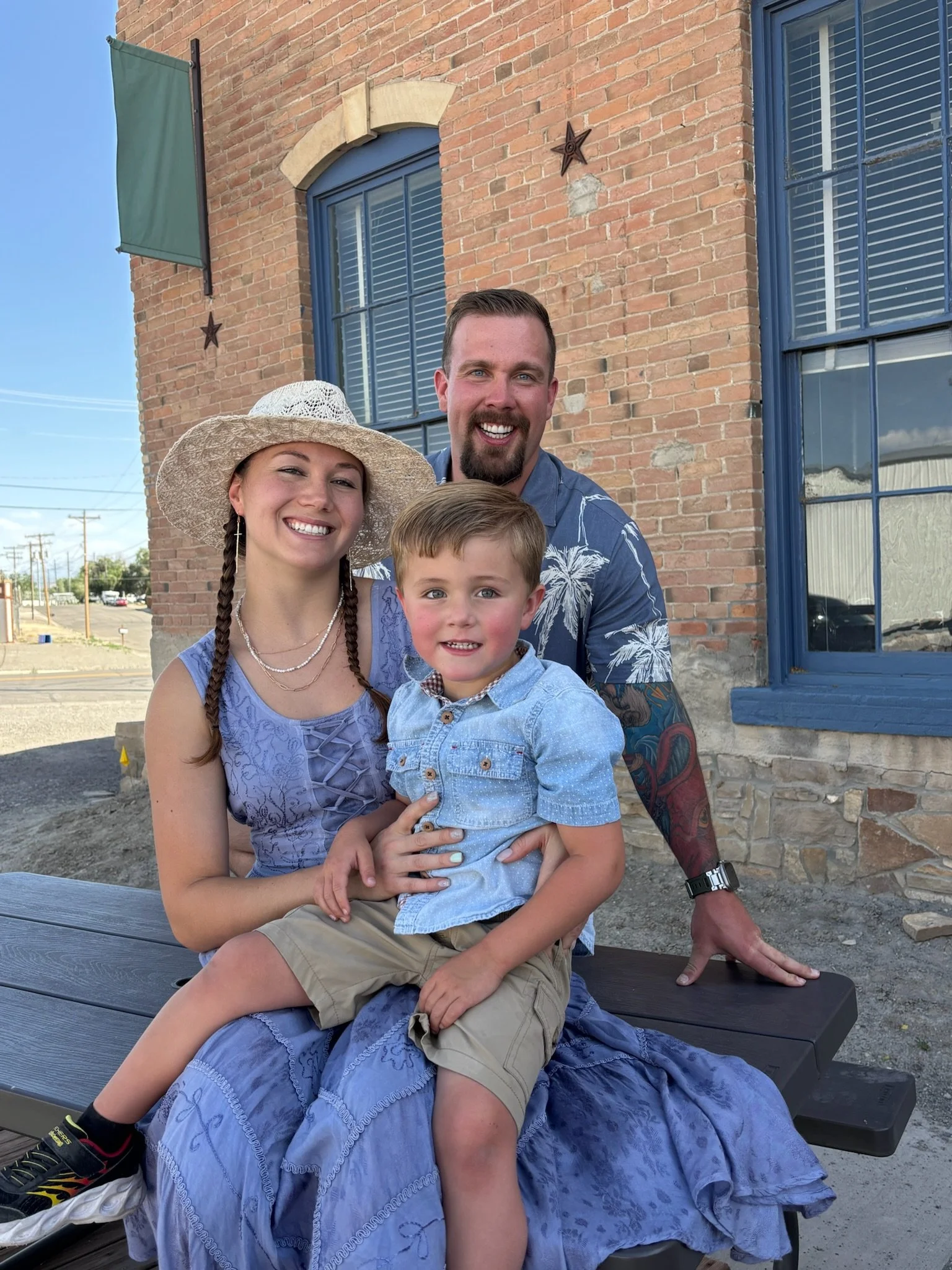 A happy family of three sitting outdoors in front of a brick building, smiling at the camera. The woman is wearing a wide-brimmed hat and a blue dress, the man is wearing a blue shirt with a tropical pattern, and the young boy is sitting on her lap, wearing a light blue shirt and khaki shorts.