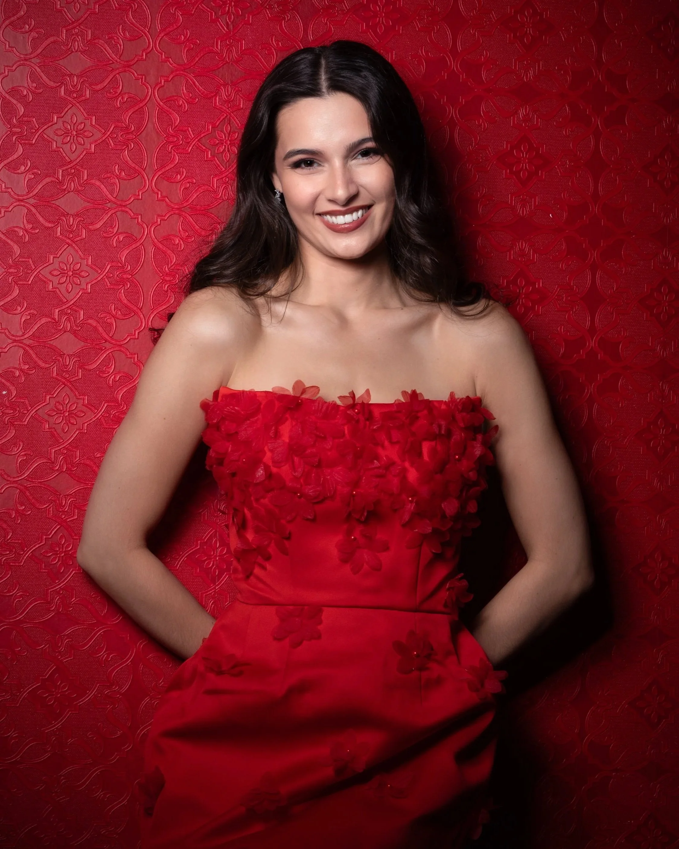 A woman with dark brown hair tucked behind her ears, wearing a strapless red dress with floral 3D embellishments on the bodice, posing against a red patterned background with a smile.