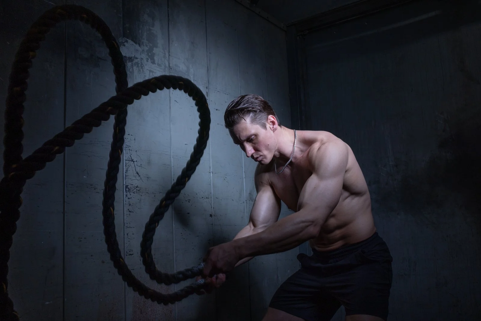 A muscular man in black shorts performs a workout with battle ropes in a dark, industrial-style gym.