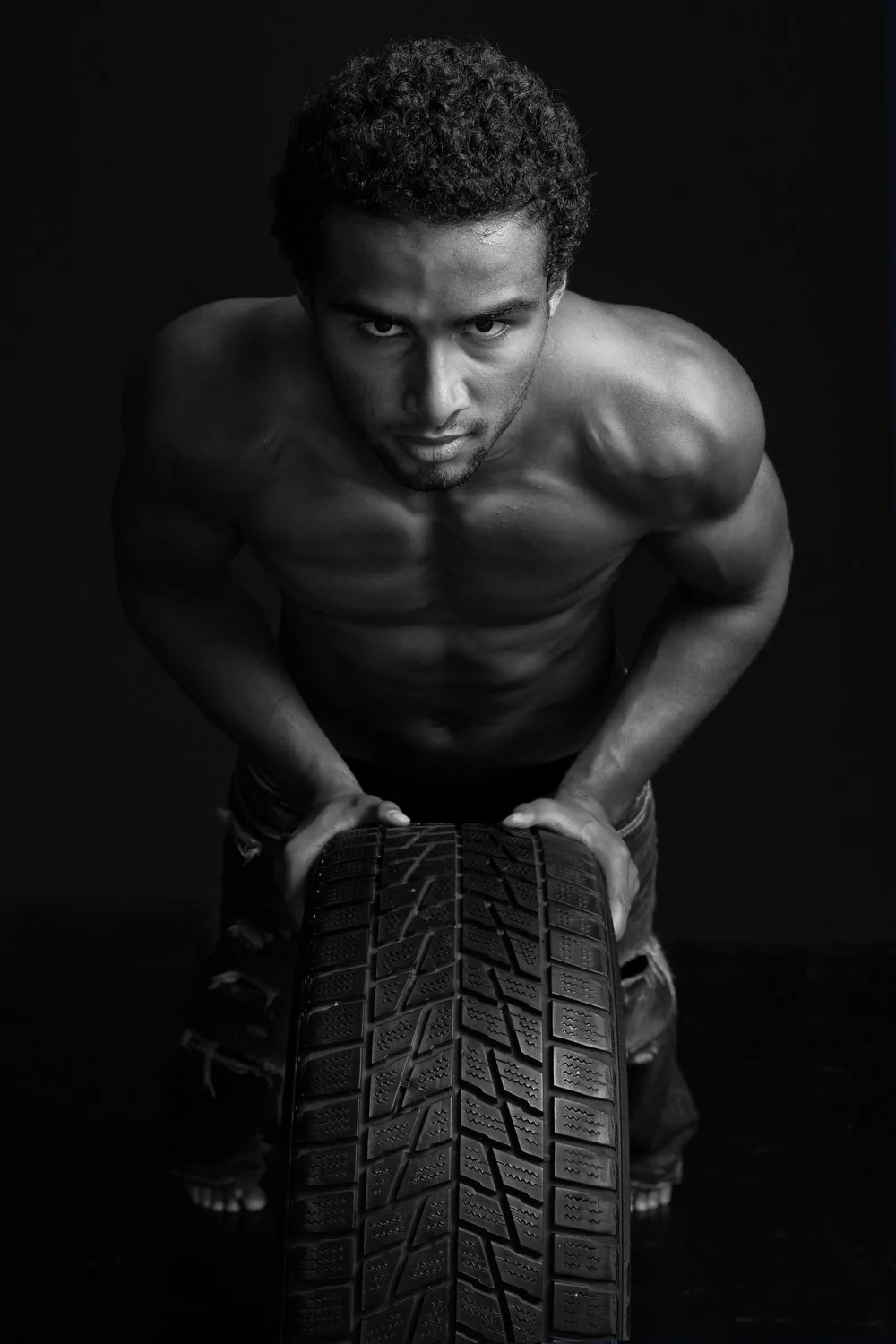 A shirtless man with curly hair and a serious expression, pushing a tire toward the camera in a dark setting.