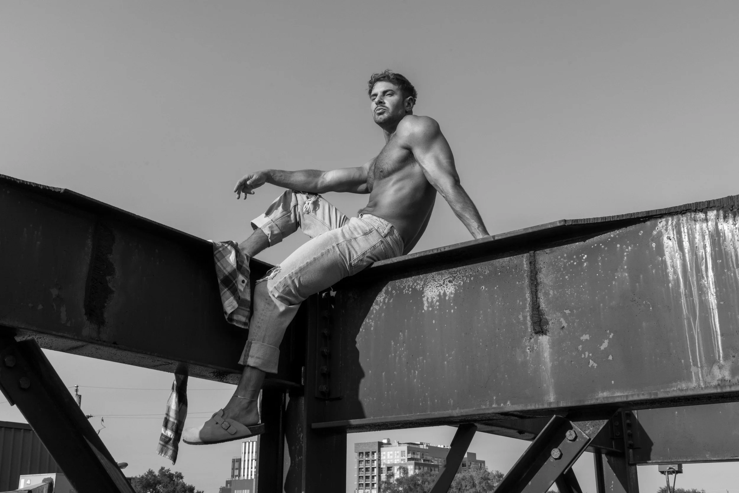 A shirtless young man with styled hair and a beard, wearing ripped jeans and slip-on sandals, sitting on a metal structure outdoors, with a city skyline in the background.