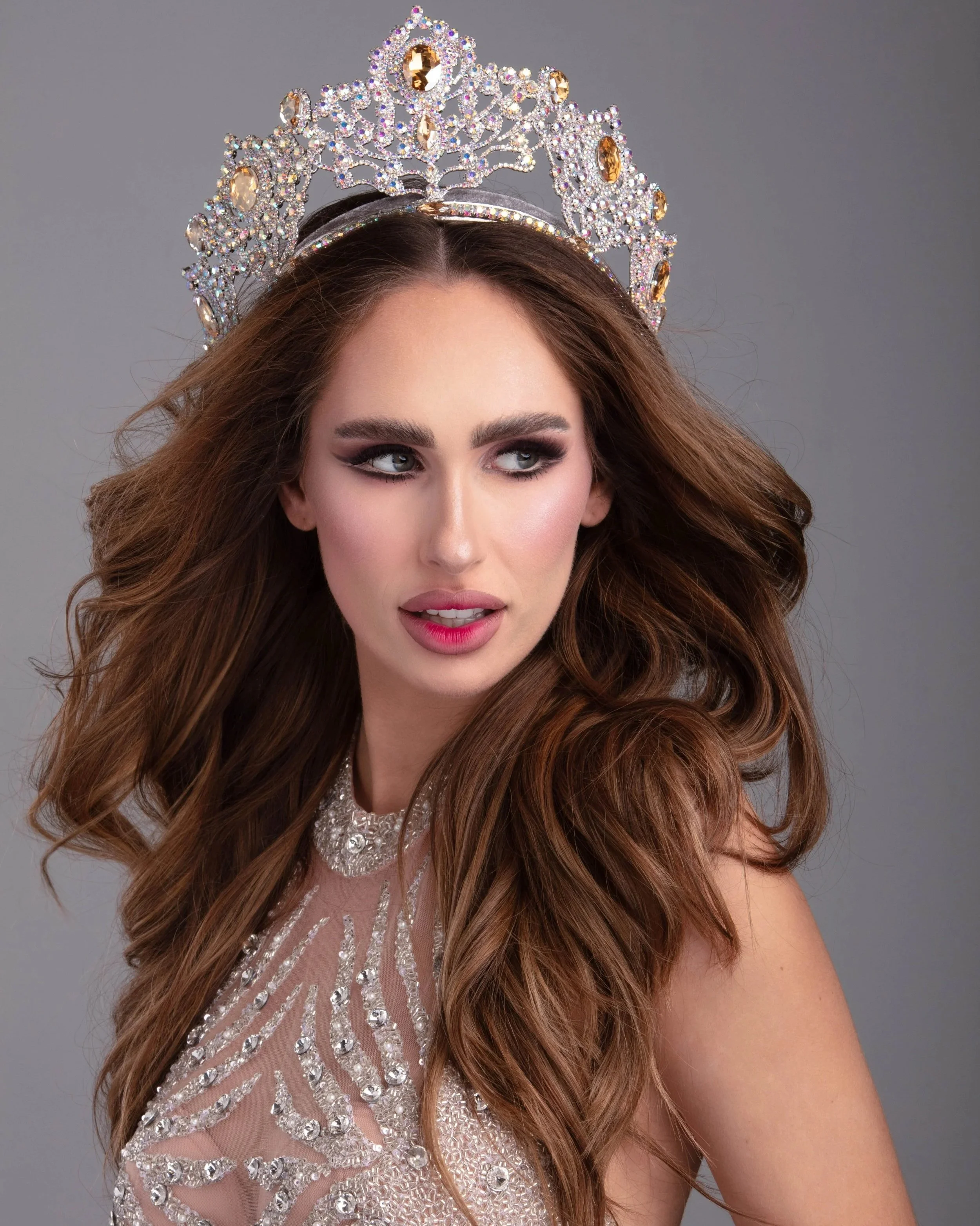 Beauty queen wearing a sparkling tiara with gemstones and an elegant, jeweled dress, looking to the side.