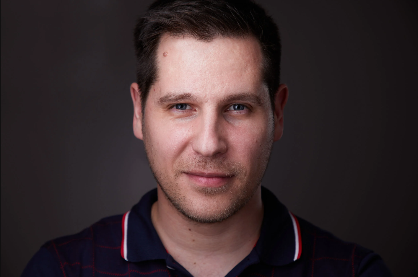 Portrait of a young man with short brown hair, blue eyes, and light facial hair, wearing a dark shirt with red and white accents, against a dark background.