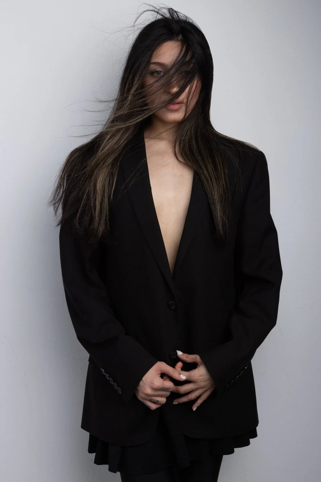 A woman with long brown hair and fair skin wearing a black blazer, with some hair covering her face, standing against a white background.
