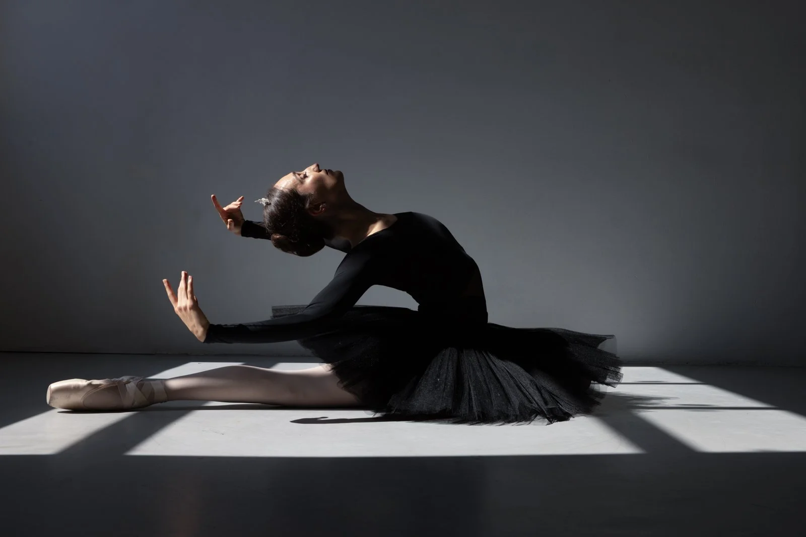 A ballet dancer in a black tutu stretches on the floor, with her head tilted back and arms raised, lit by dramatic lighting creating shadows.