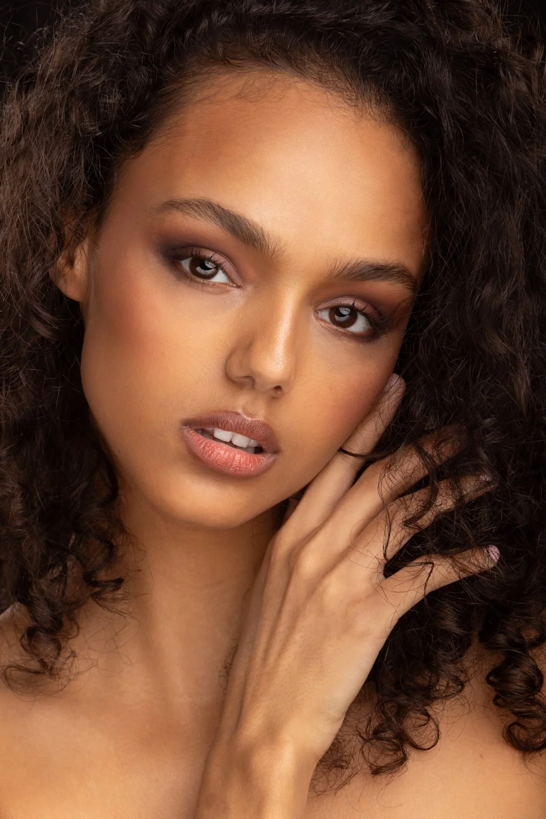 A close-up portrait of a woman with curly hair and natural makeup, touching her face gently with her hand.