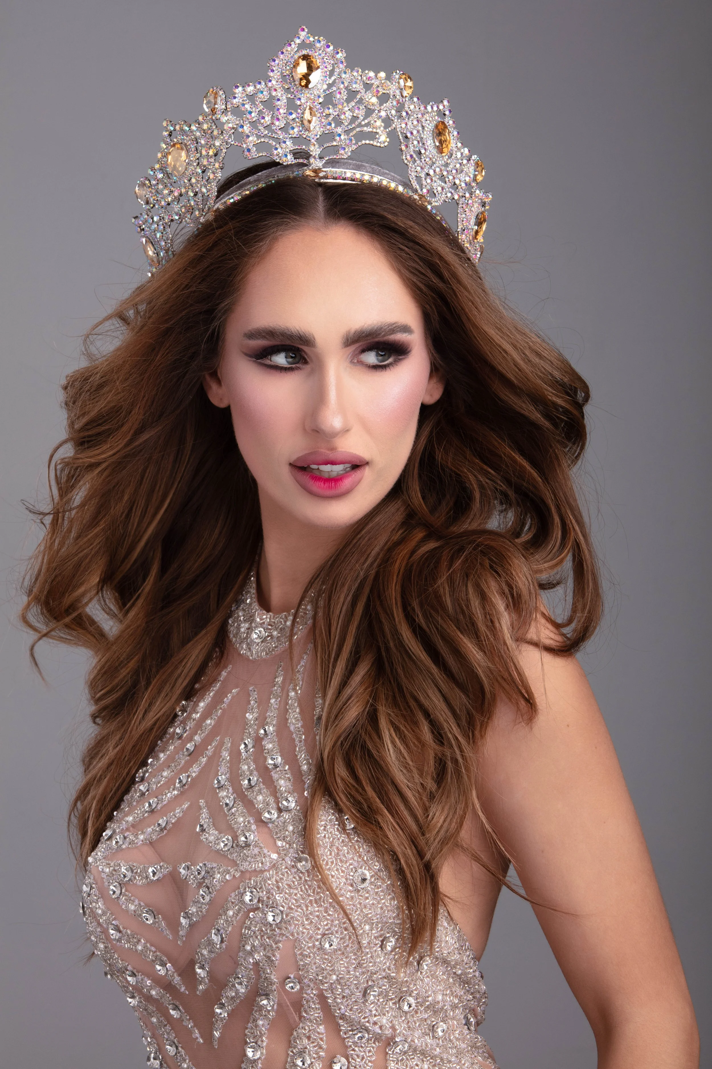 A woman with long, wavy brown hair wearing a rhinestone crown, a sheer gown with intricate rhinestone and bead embellishments, and makeup, posing against a gray background.