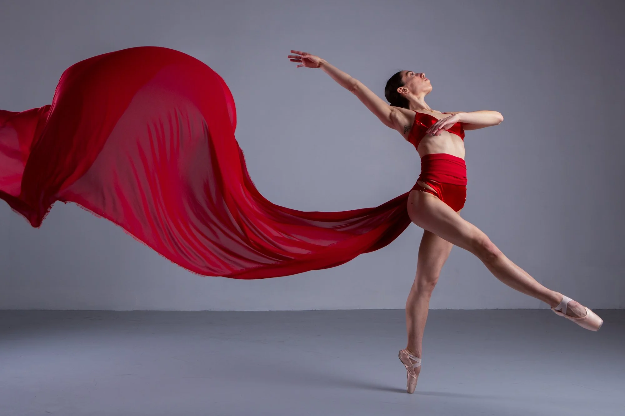 A female ballet dancer in a red outfit performing a pose on pointe, with a flowing red fabric extending behind her against a plain gray background.