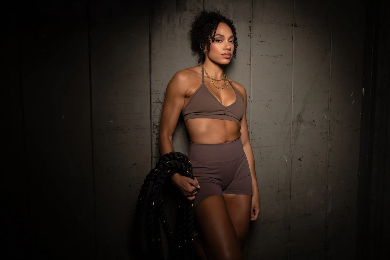 A woman wearing a sports bra and shorts, holding a thick black rope, standing against a dark wall.