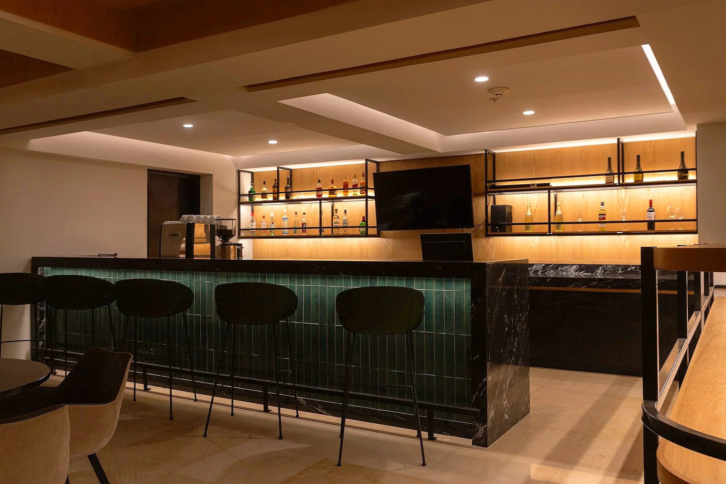 Modern bar interior with black bar counter, high black chairs, wooden shelving with liquor bottles, a TV, and warm lighting.