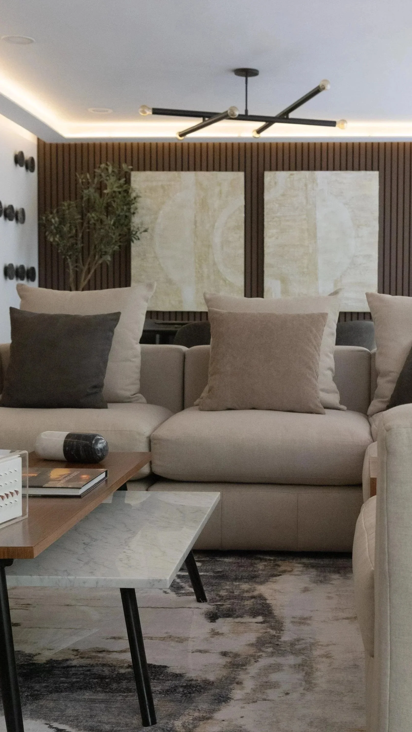 Modern living room with a beige sectional sofa, black and beige throw pillows, a wooden coffee table with a white marble top, decorative items, and contemporary wall art featuring abstract designs and wall-mounted décor.