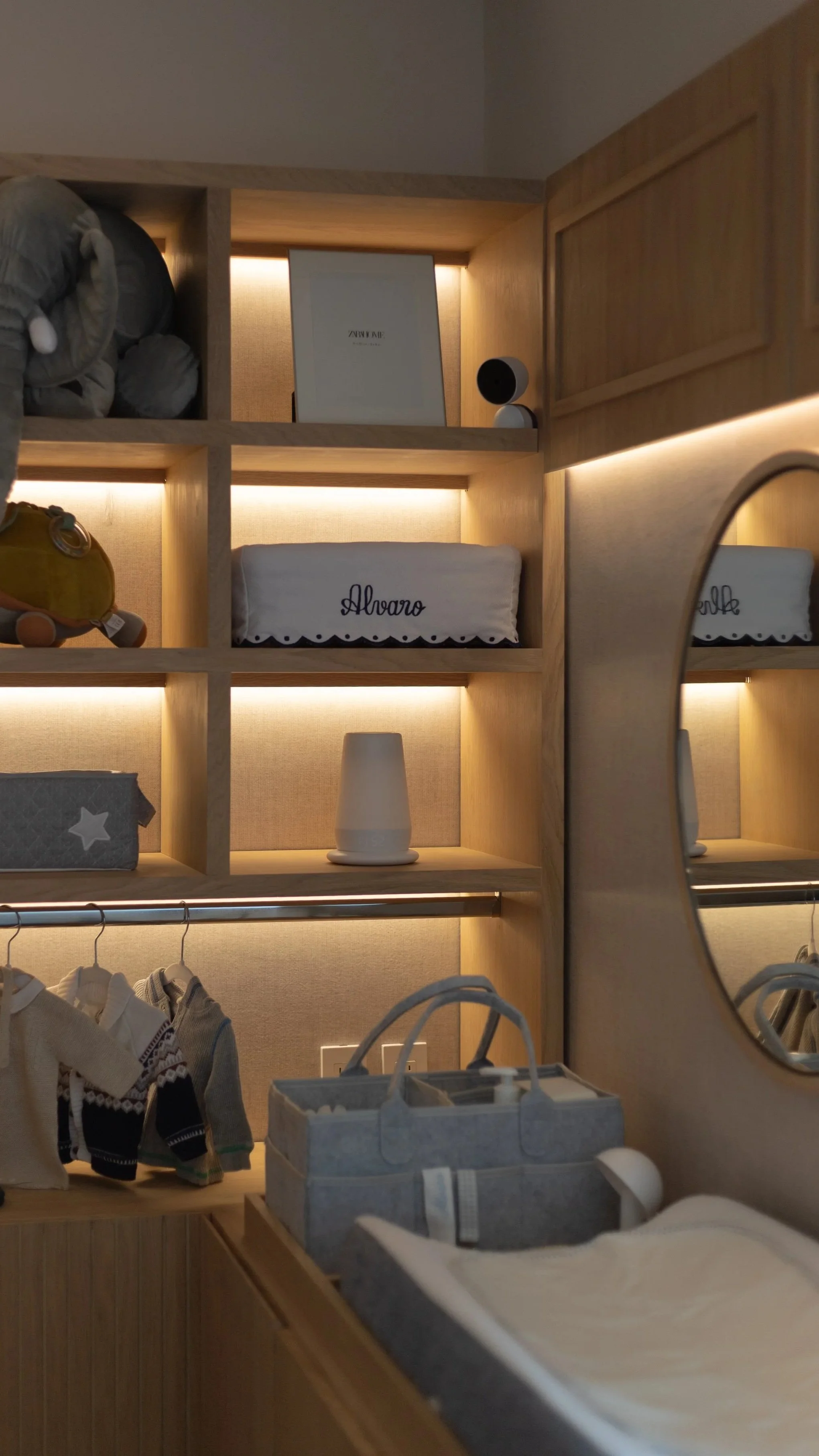 Estantería de madera con objetos decorativos y ropa de bebé, incluyendo una caja de pañales y un espejo redondo.