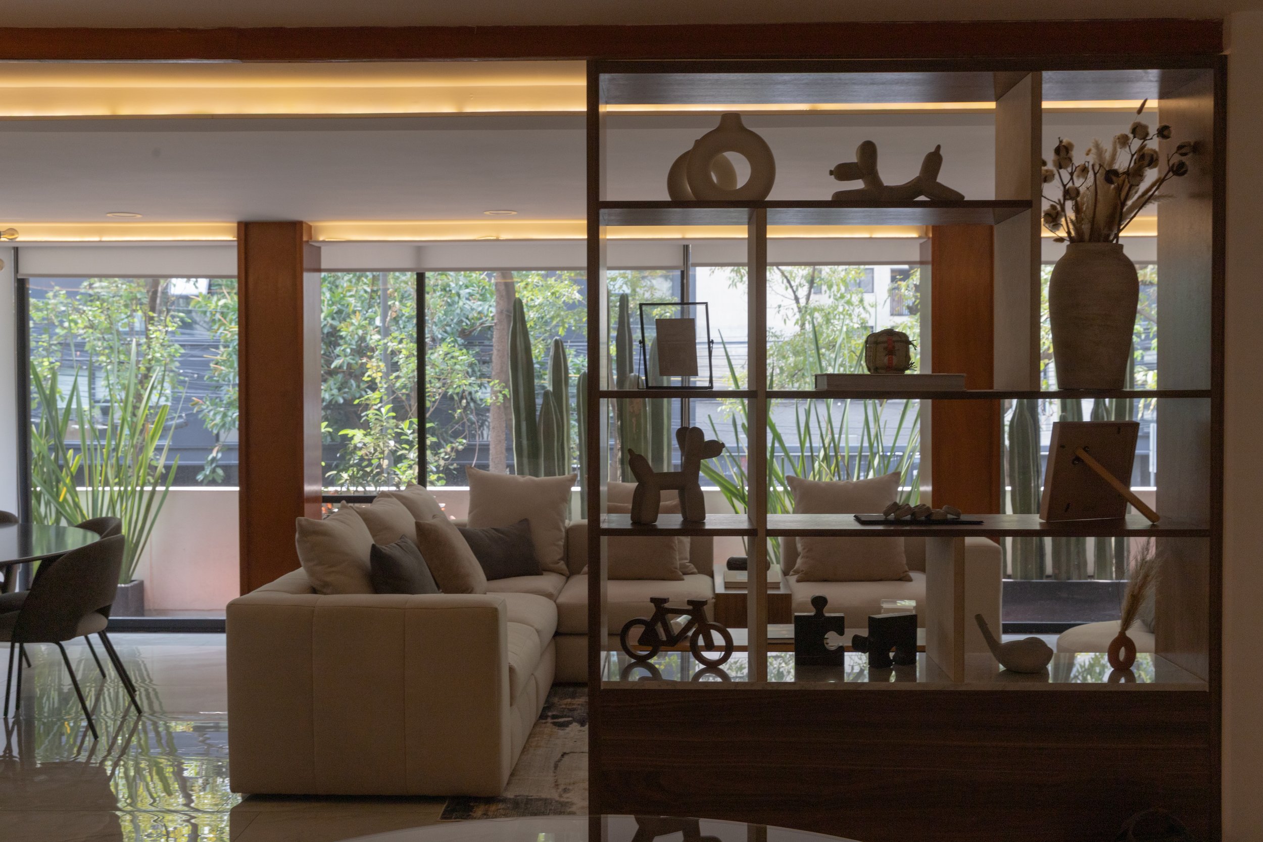 Sala de estar con sofá beige, cojines en tonos neutros, estantería de madera con decoraciones y plantas, ventanas grandes con vista a árboles y plantas exteriores.