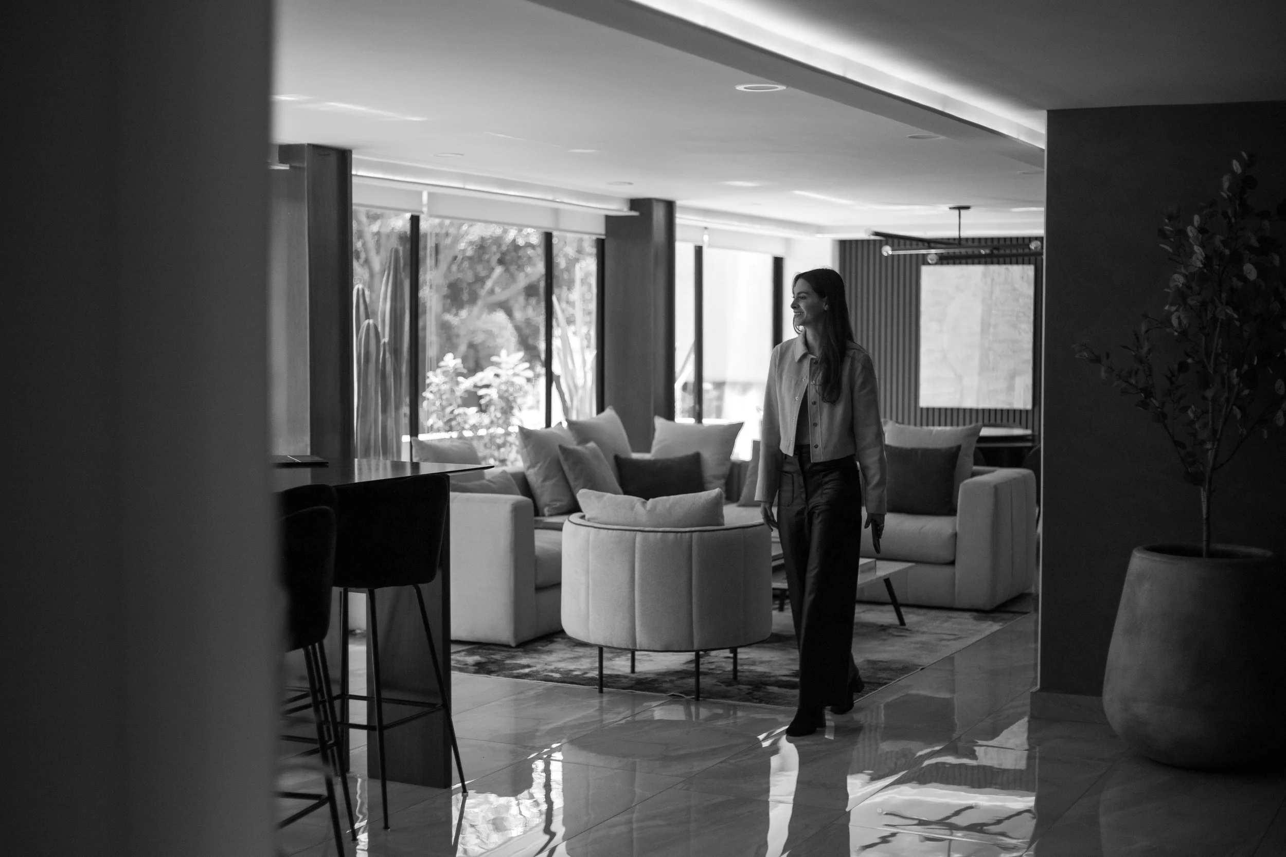 A woman walking through a modern living room with large windows and contemporary furniture, including multiple sofas and chairs, in black and white.