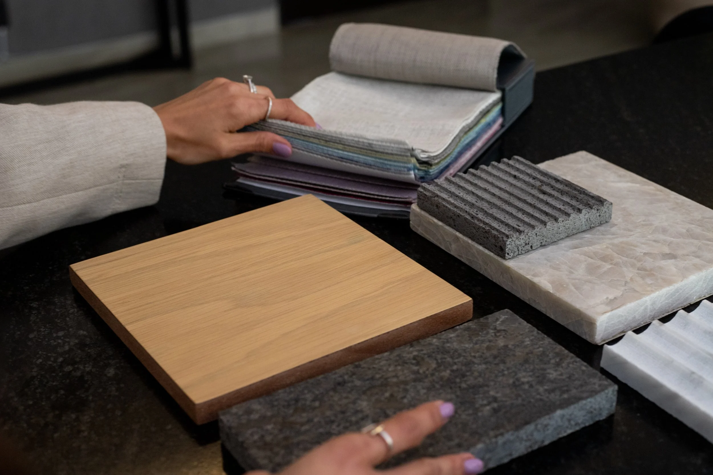 Muestra una mesa de trabajo con muestras de diferentes materiales como madera, piedra y concreto, y una persona con uñas pintadas revisando una libreta de muestras de colores y texturas.