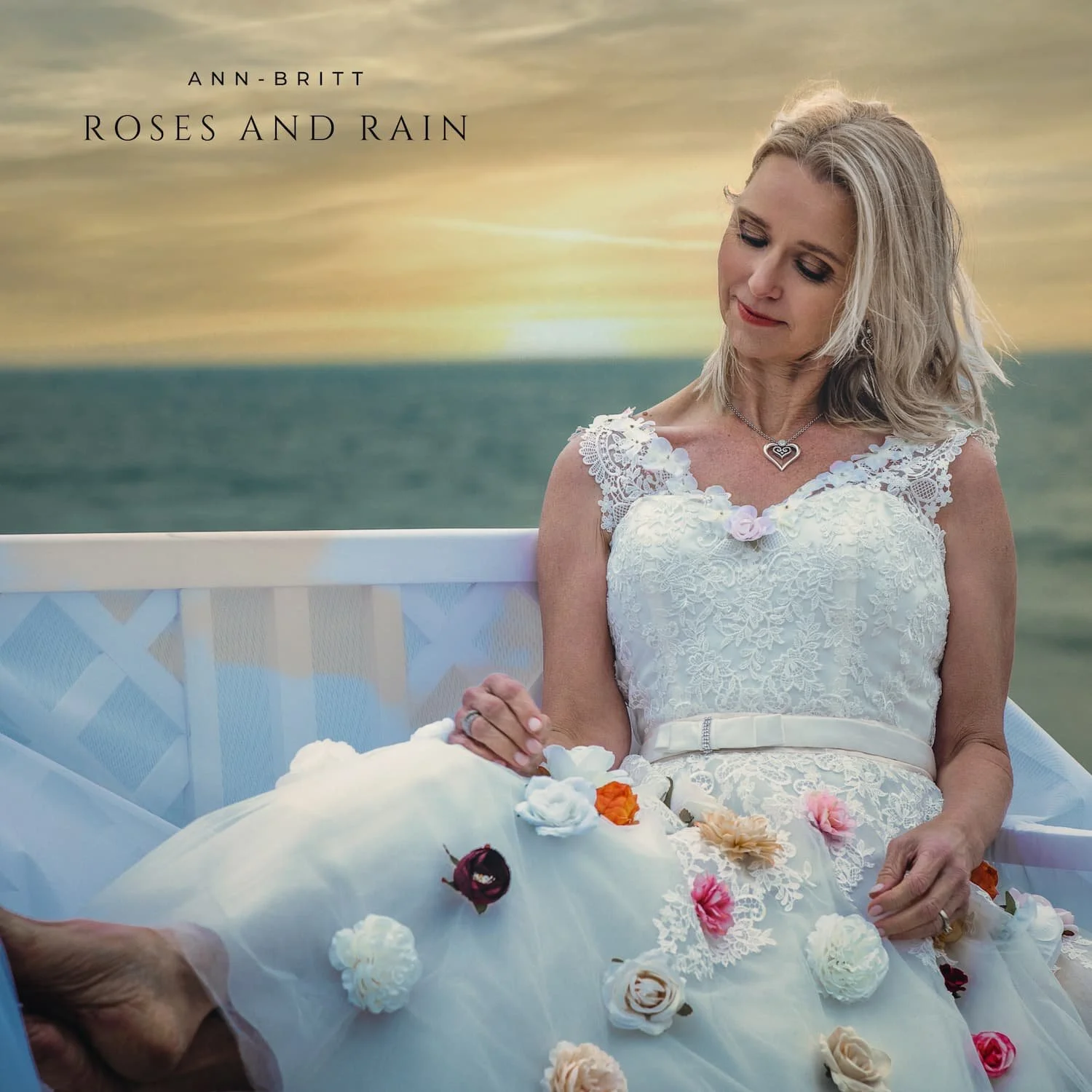 A woman in a white lace dress with a heart-shaped necklace holding hands with someone during a wedding or celebration, with a sunset sky and ocean in the background.