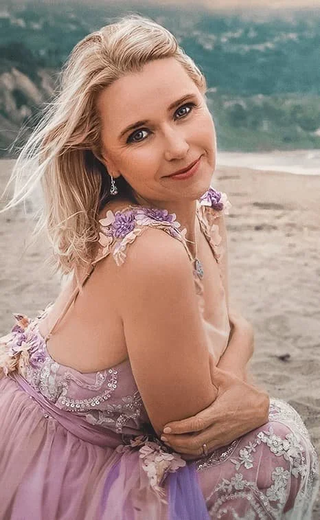 A woman with blonde hair sitting on a sandy beach, wearing a pink floral dress, with a gentle smile and a scenic ocean background.