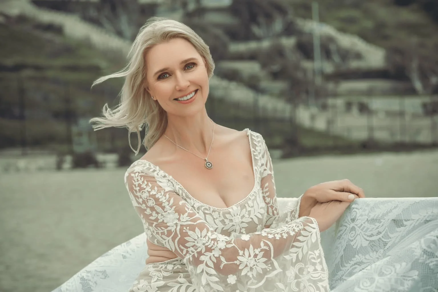 A woman with blonde hair smiling and wearing a lace dress with floral patterns, standing outdoors near water with rocky hills in the background.