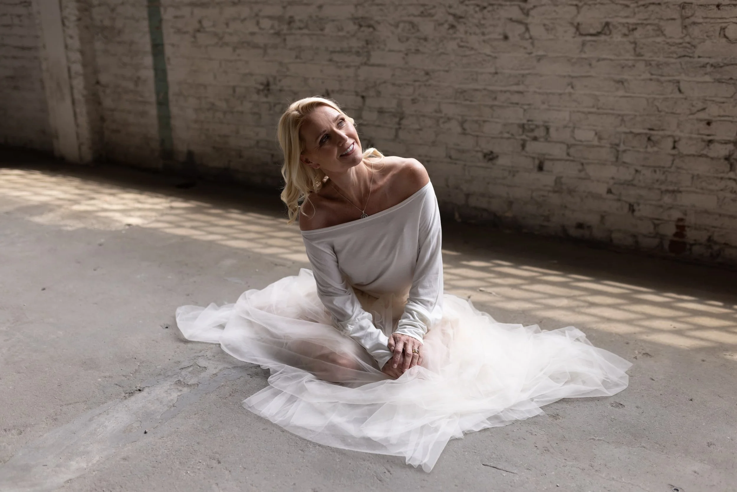 A woman in a white off-shoulder dress sits on the ground in front of a brick wall, smiling and looking up, with sunlight casting shadows on the wall.