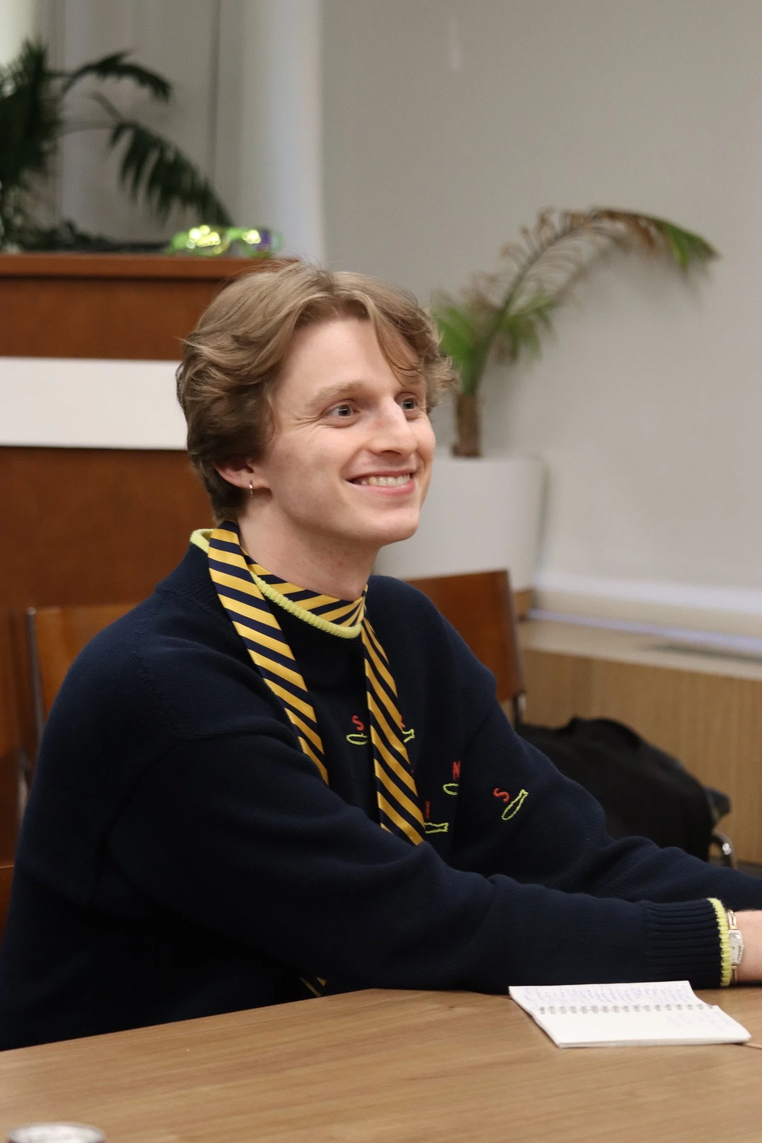 Image of Owen Boyce as "JOSH" in The Antipodes. He is wearing a navy blue turtleneck with a blue and yellow tie around his neck like a scarf. He is smiling and sitting in an office setting.