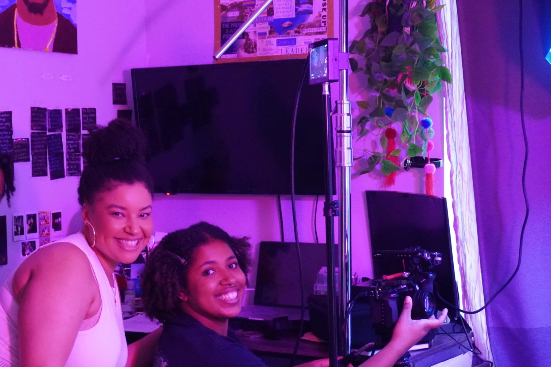 women smiling and posing for a photo indoors with purple lighting. One is wearing a white tank top, and the other two are wearing dark-colored tops. There are computer monitors, a DJ controller, a large TV screen, a potted plant, and wall decor in the background.