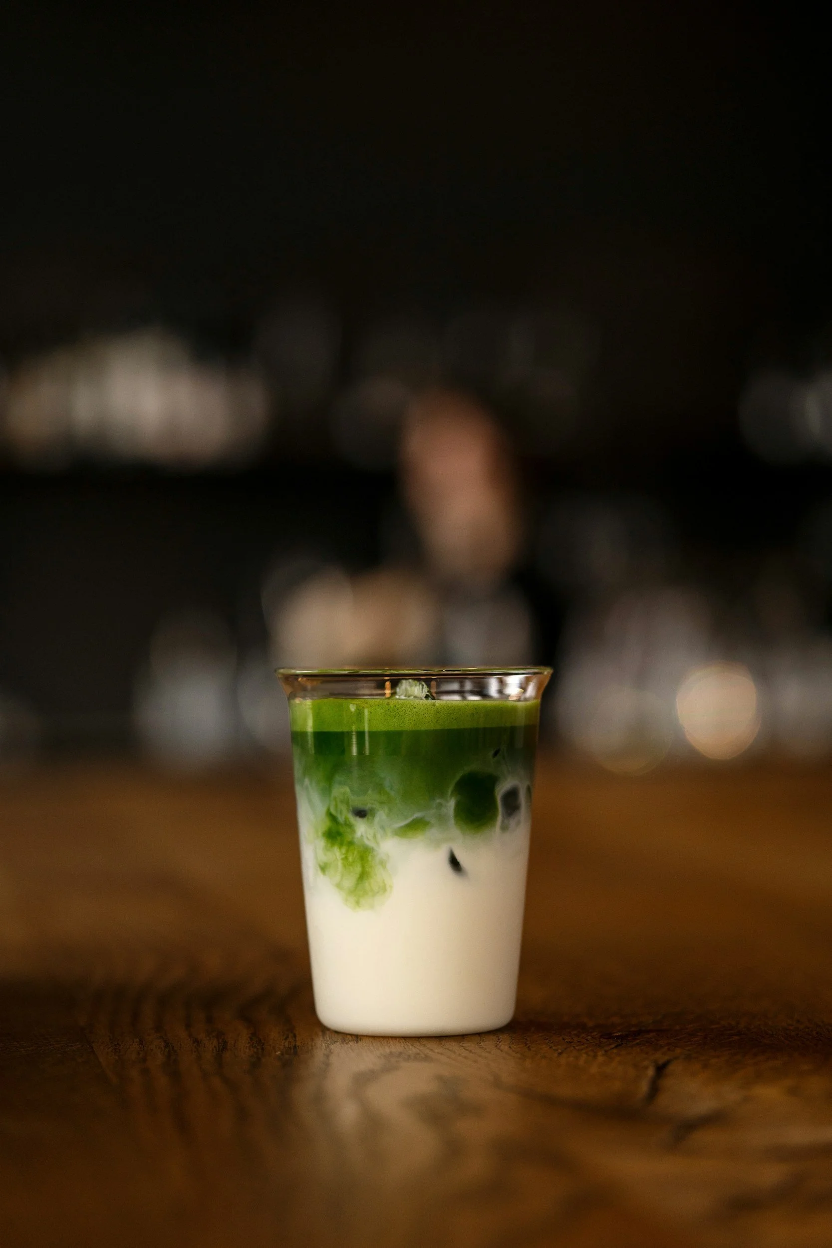 A tall glass of a layered matcha latte with green tea, milk, and black tapioca pearls on a wooden surface, with a dark blurred cityscape in the background.
