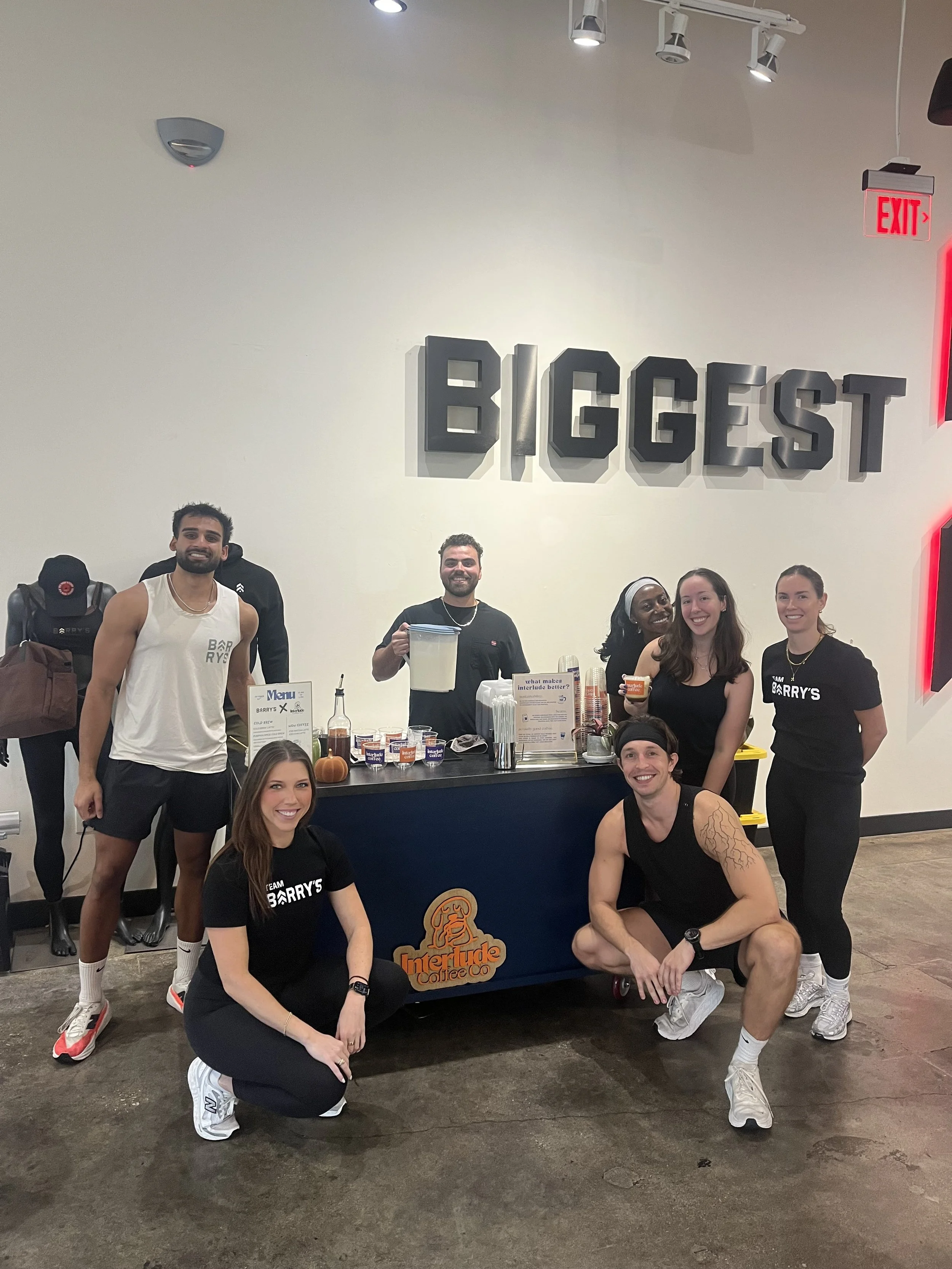 Group of seven people at a coffee shop, some standing and some kneeling in front of a counter with the logo "Interlude Coffee"; a menu and various coffee supplies are visible on the counter.