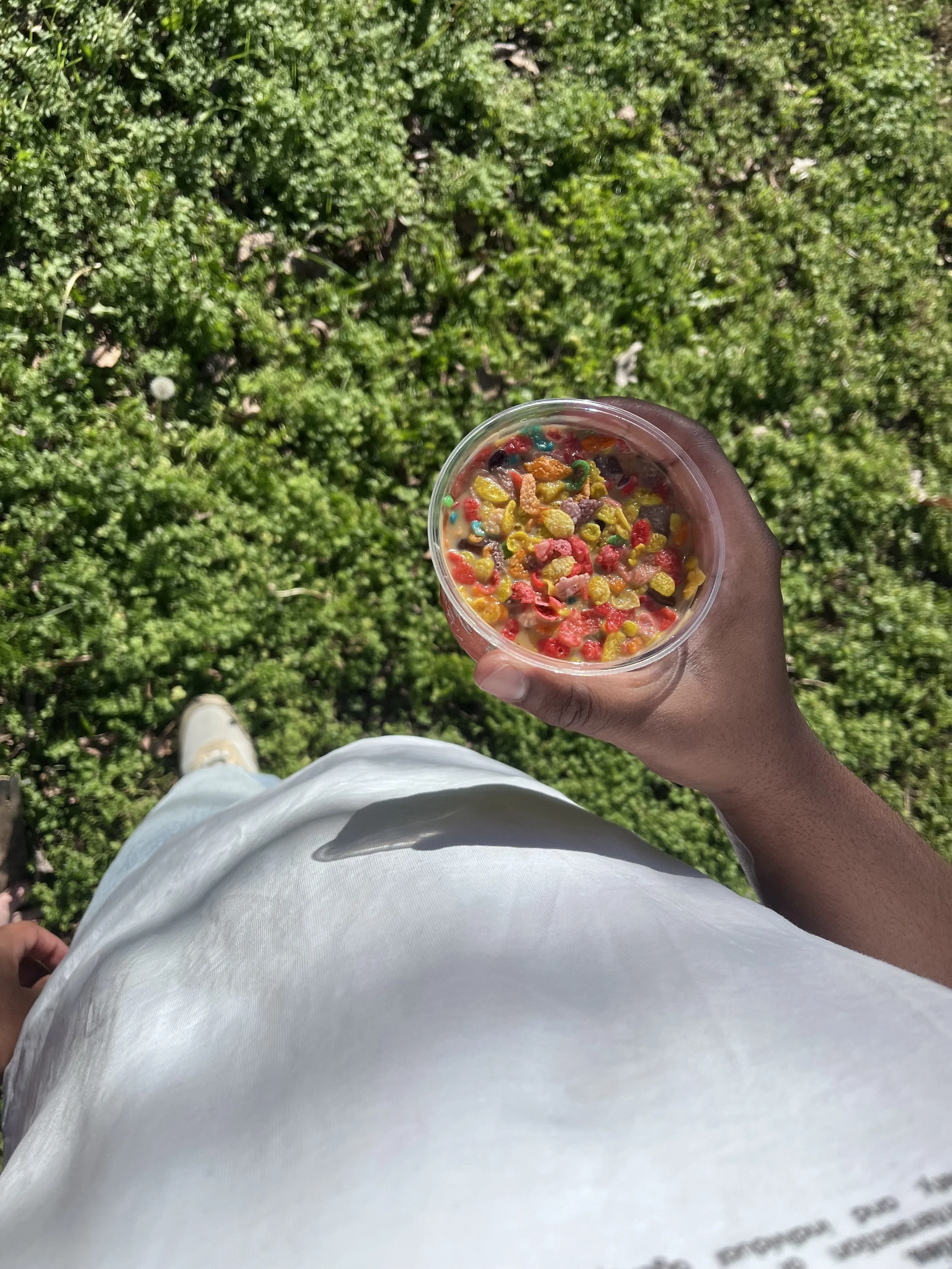 A person holding a clear plastic cup filled with colorful breakfast cereal and milk, standing outdoors on a grassy area, with a view of their white clothing and beige shoes.