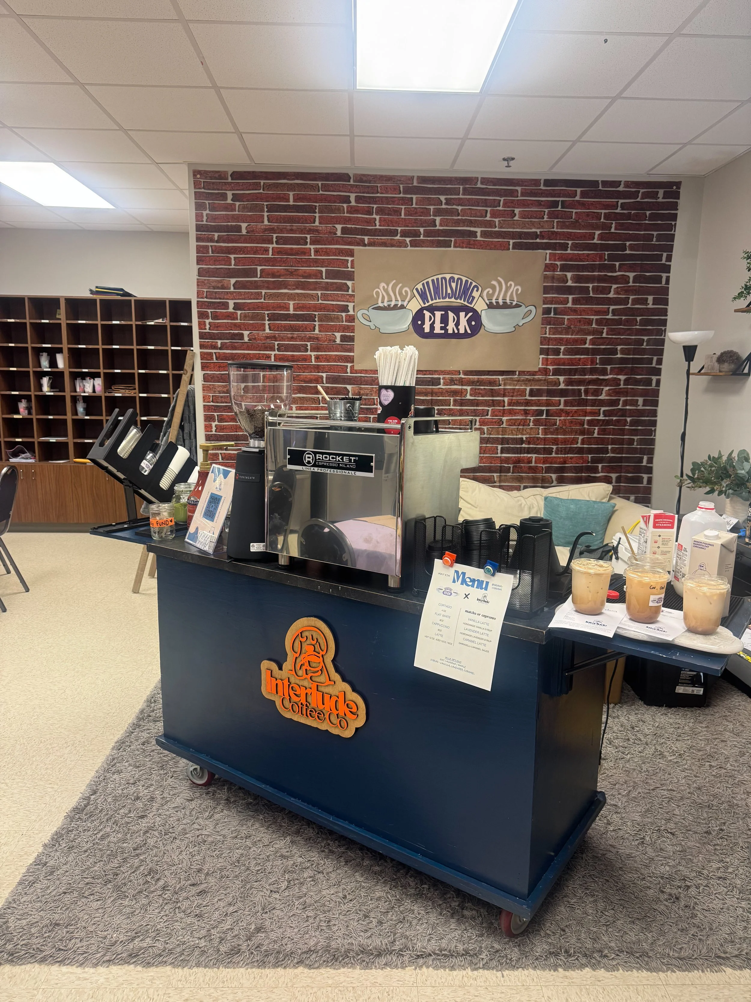 Coffee counter with a red brick wall and a sign that reads 'Windsong Perk' with steaming coffee cups. There is a coffee machine, cups, and drink containers on the counter, with a menu sign hanging. To the left, there are open cubby shelves with supplies, and the floor is carpeted in front of the counter.