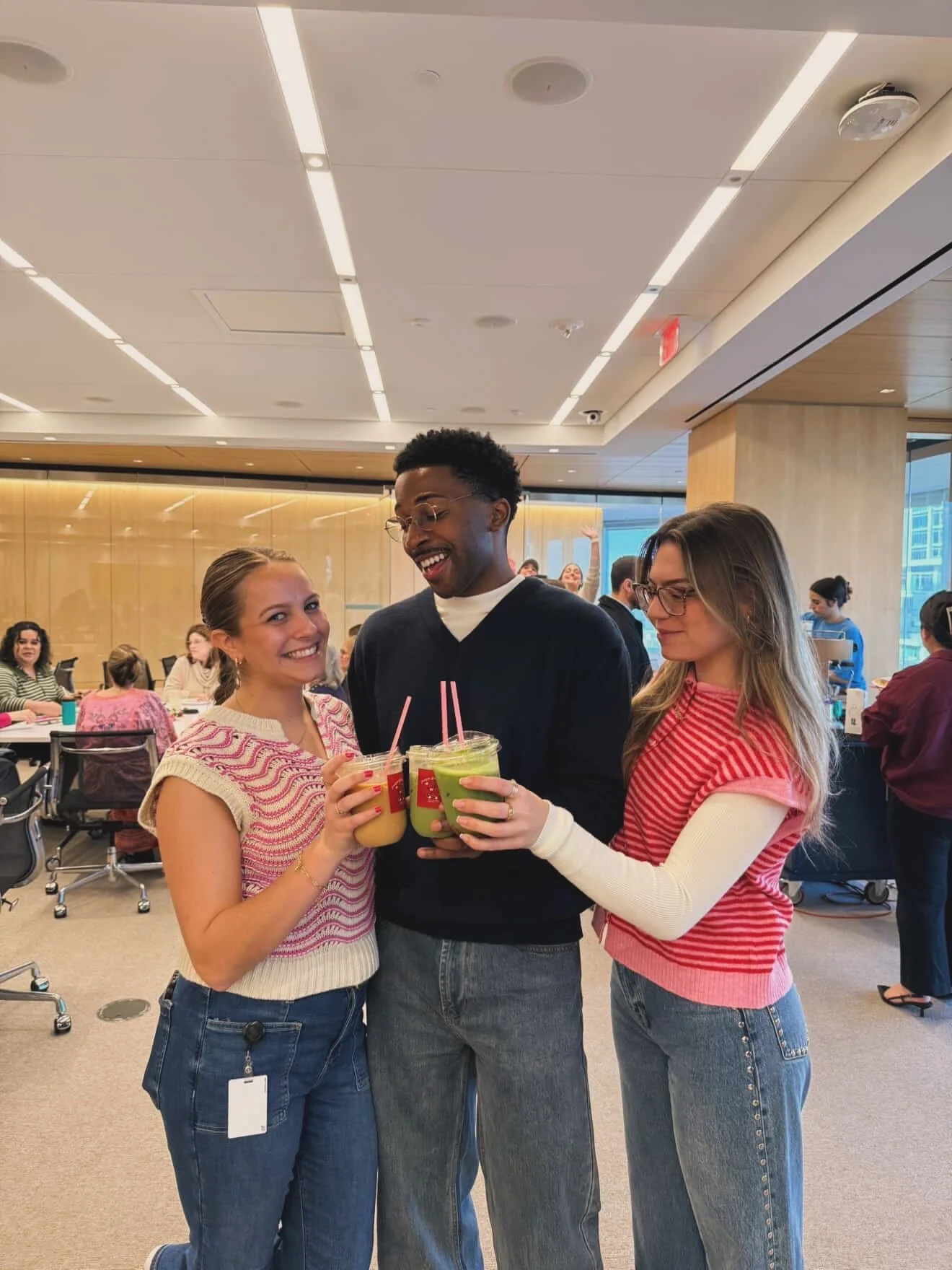 Three friends smiling and holding smoothies in a modern, busy cafe or restaurant with other people in the background.