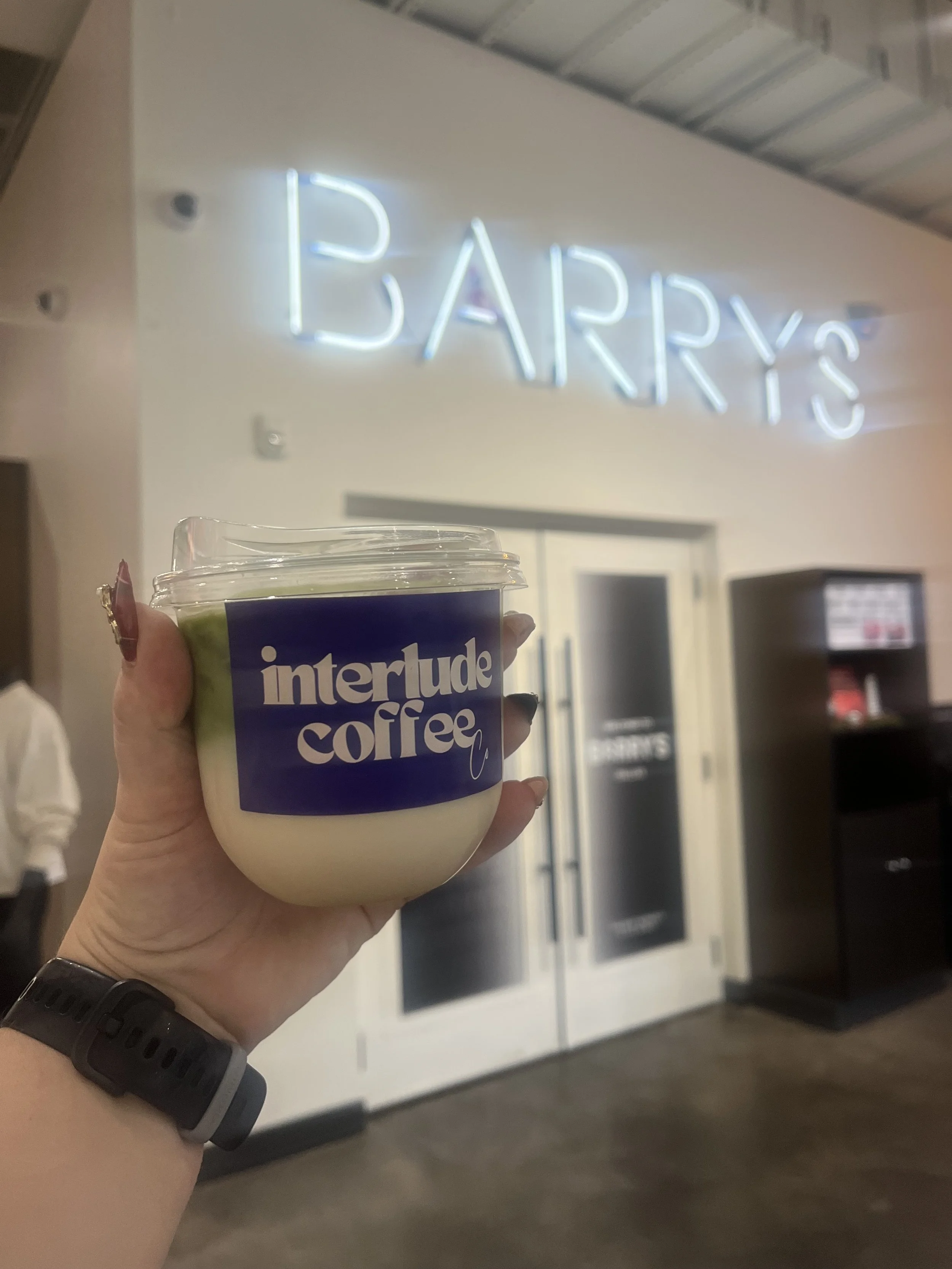 A person holding a cup of iced coffee with a purple and white logo that says 'interlude coffee' in front of a neon sign that says 'EARRYS' in a shopping mall or cafe interior.