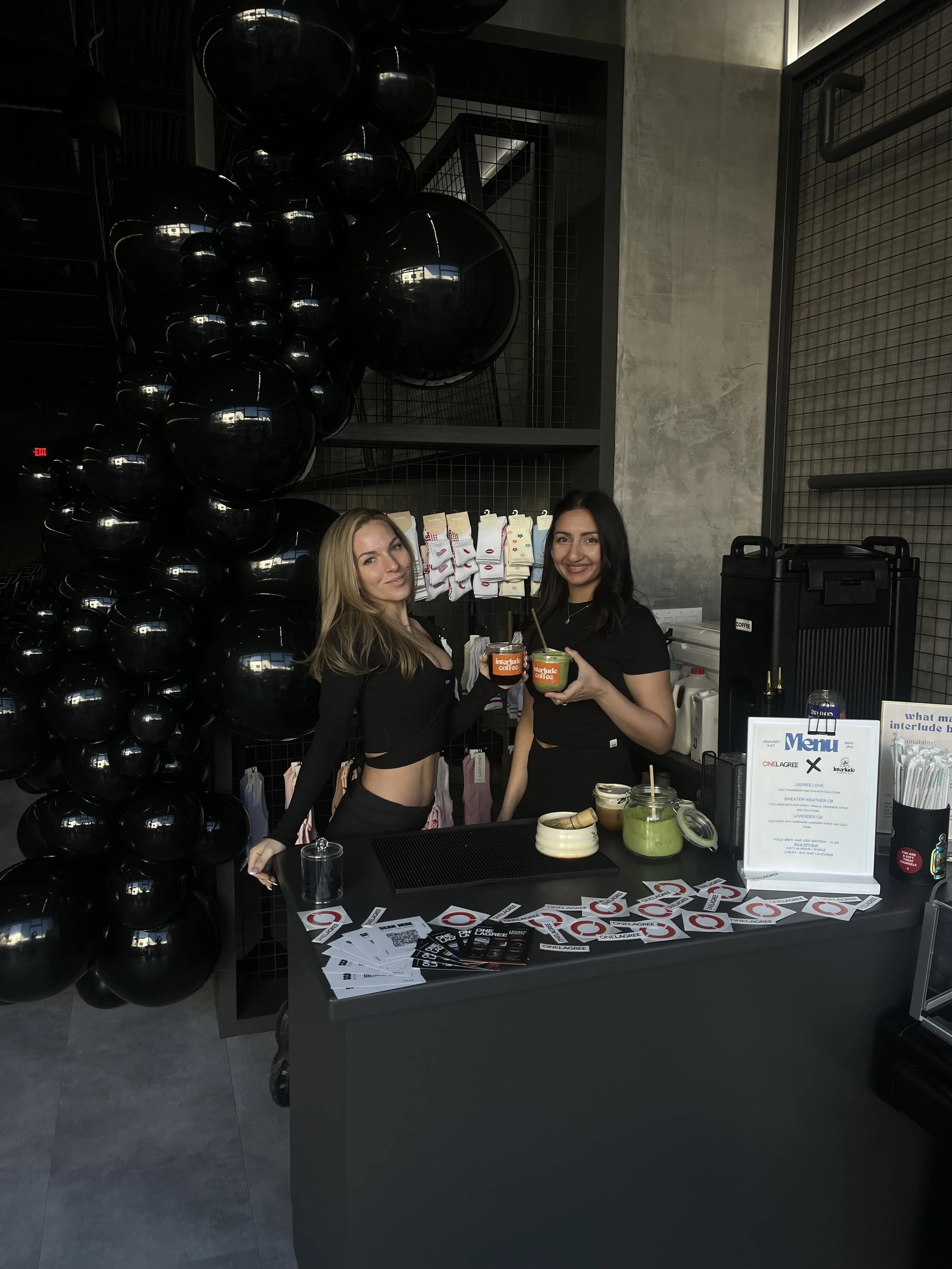 Two women standing behind a counter at a coffee shop, holding cups of coffee and smiling. The counter has flyers, a menu, and jars of drinks. Black balloons decorate the background, and there are various coffee drinks and supplies on the counter.