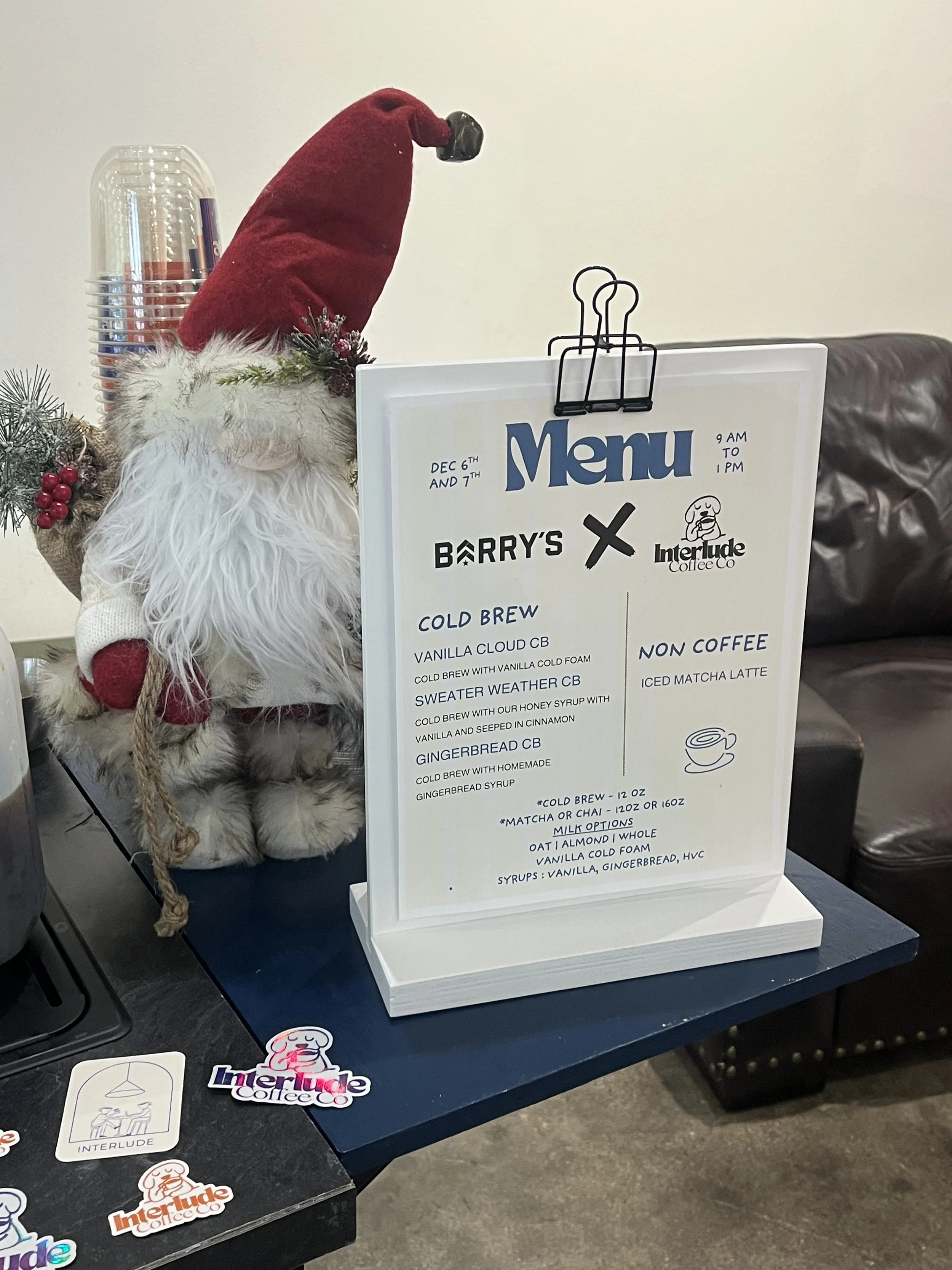 A decorative Santa Claus figure with a red hat, a white beard, and a white fluffy suit, standing next to a menu sign on a table, with a small Christmas plant and some stickers nearby.