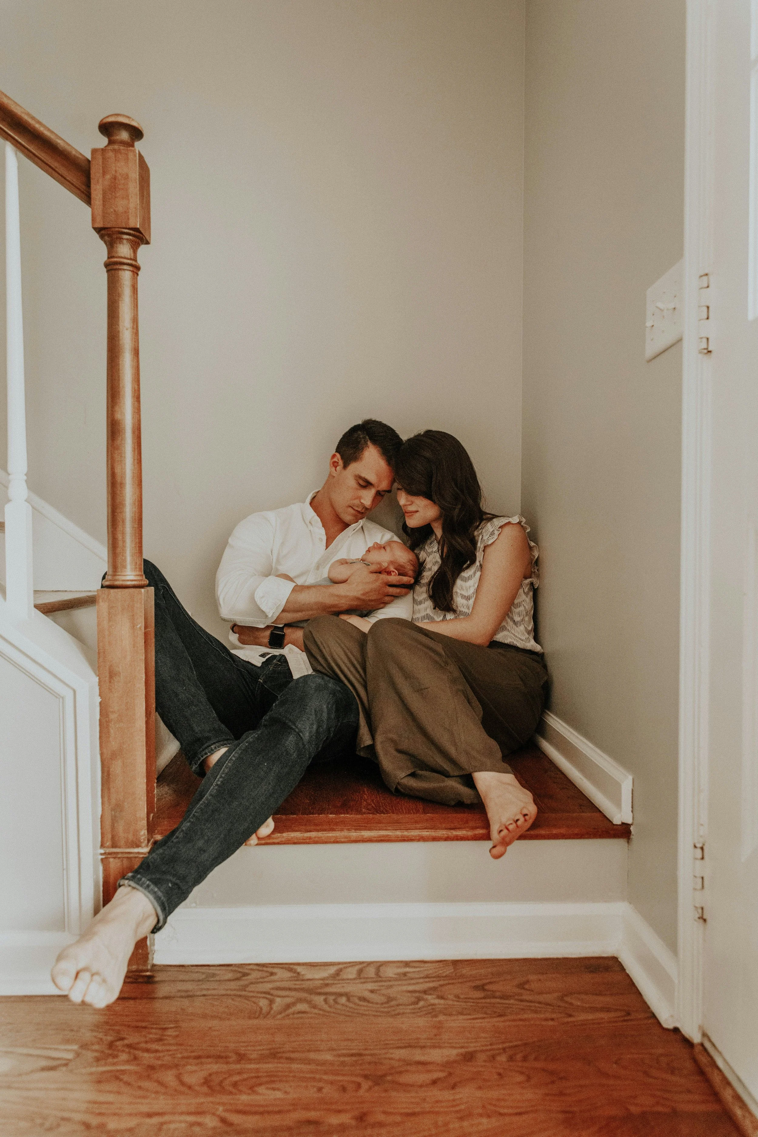 A family of three sitting on a wooden staircase, with the father holding a newborn baby, and the mother sitting close, all appearing happy and intimate.