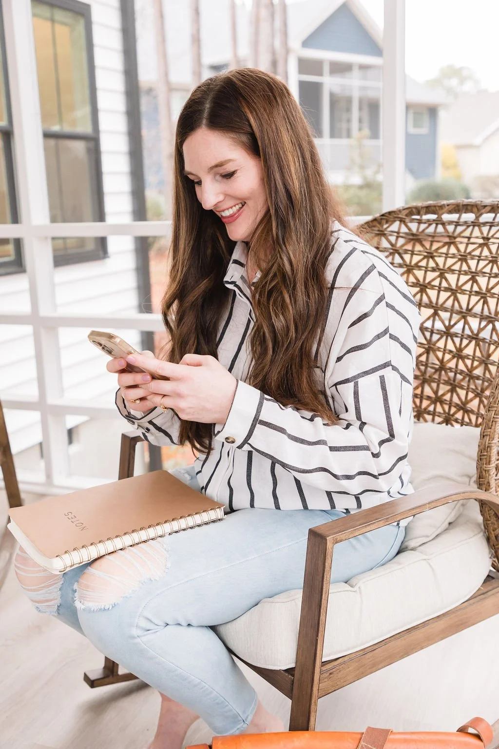 A woman with long brown hair sitting on a cushioned wooden chair, smiling while looking at her phone. She is wearing a white and black striped shirt and light blue ripped jeans, with a notebook or planner on her lap. The setting appears to be a bright porch or sunroom with windows and neighboring houses visible outside.