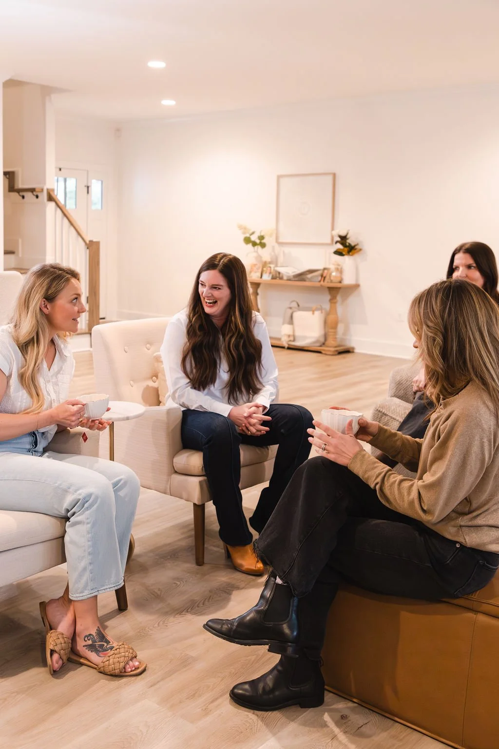 Group of five women sitting in a cozy living room, engaging in conversation and smiling, with some holding cups.