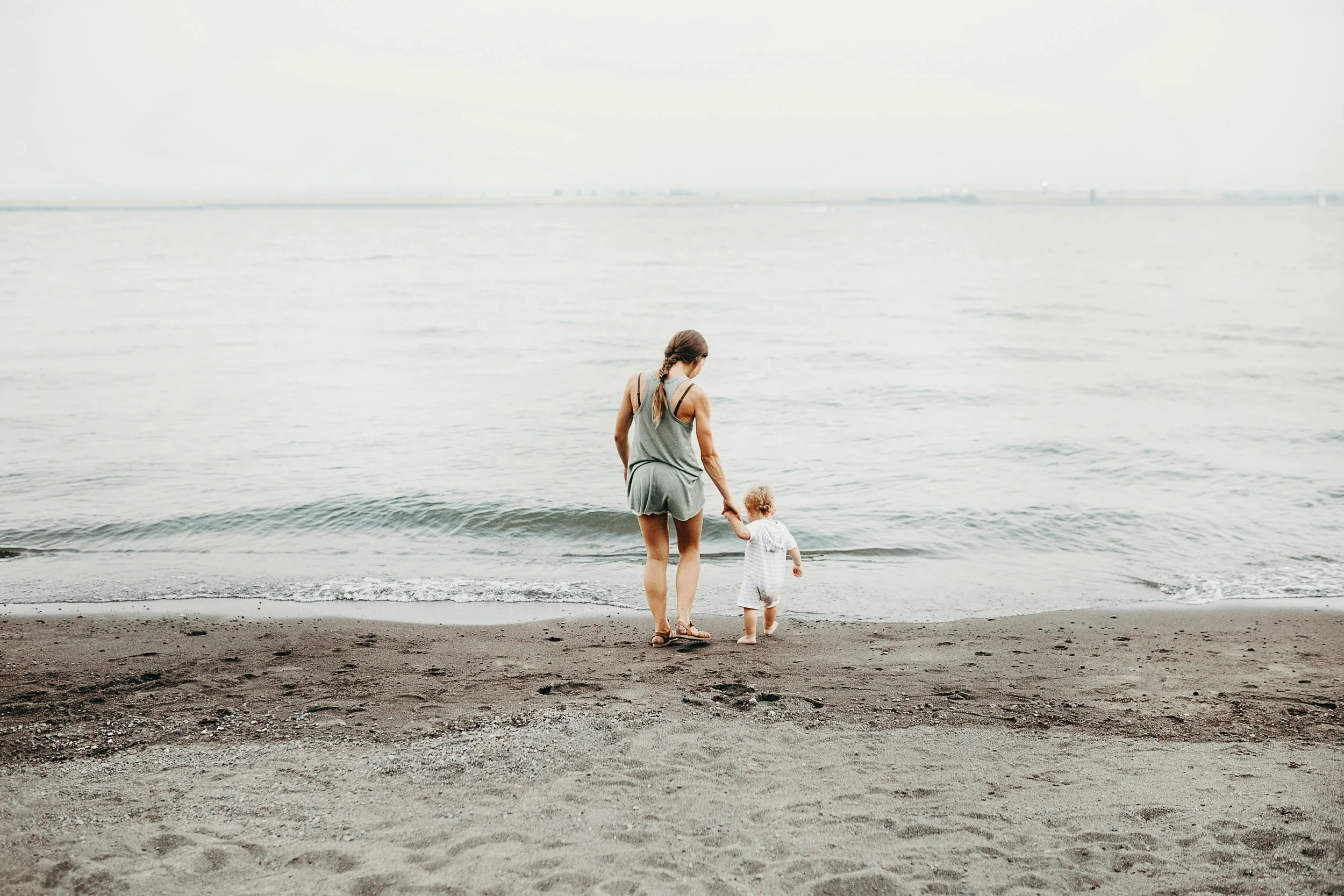 A woman and a young child walking hand in hand along the shoreline of a beach, with calm water in the background.