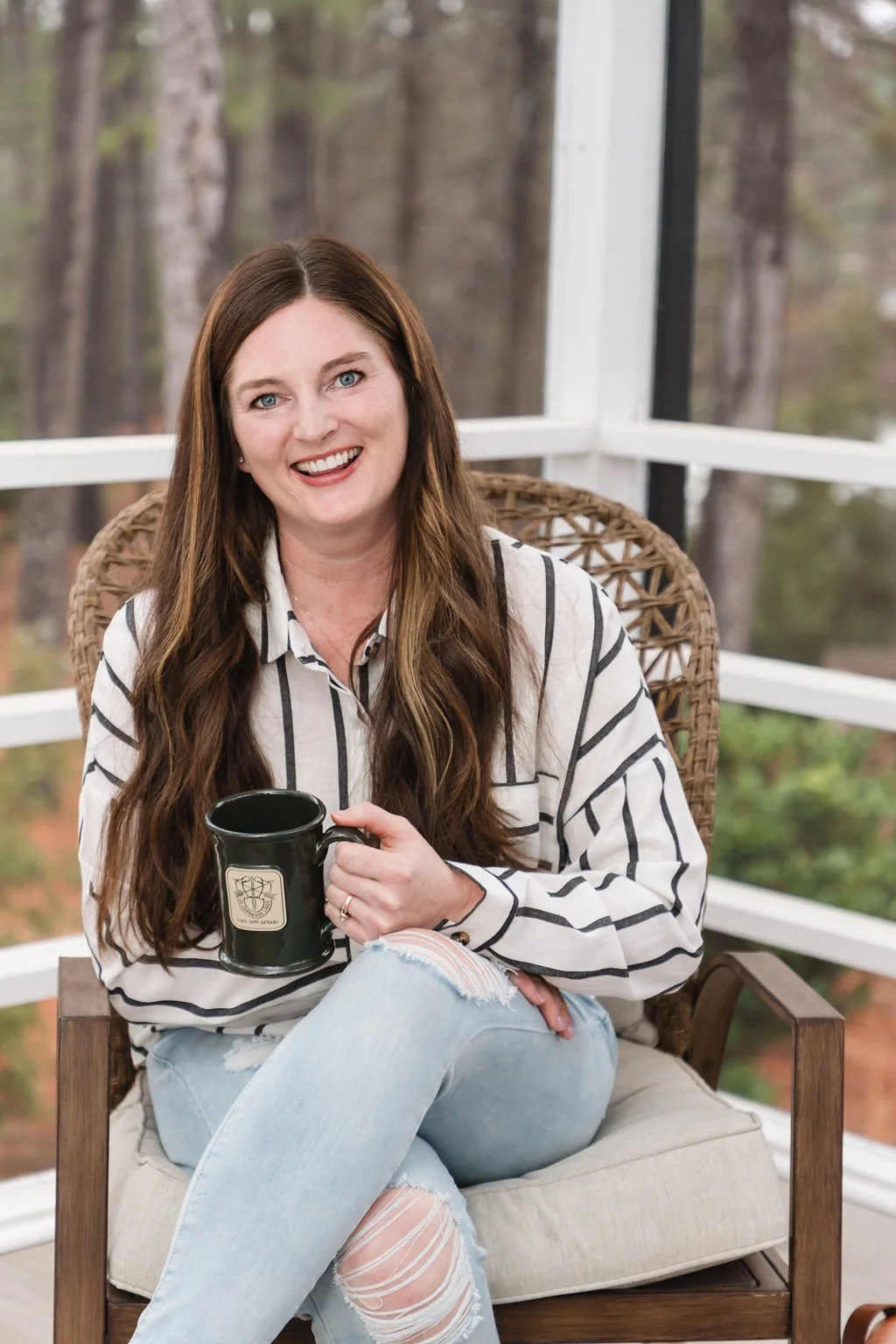 Holistic Health Founder, Breanna Imperial sitting on a wicker chair on a screened porch, holding a black mug, smiling at the camera, with a wooded background outside.