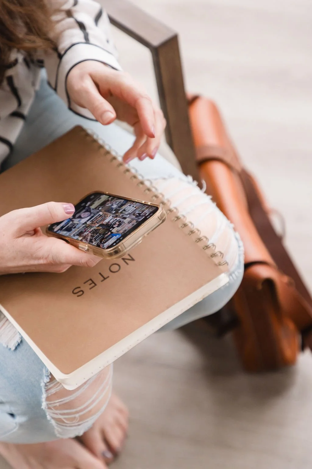 Person sitting on a wooden bench, holding a smartphone and a spiral notebook with the word 'NOTES' on the cover. The person is wearing ripped jeans and a striped long-sleeve shirt.